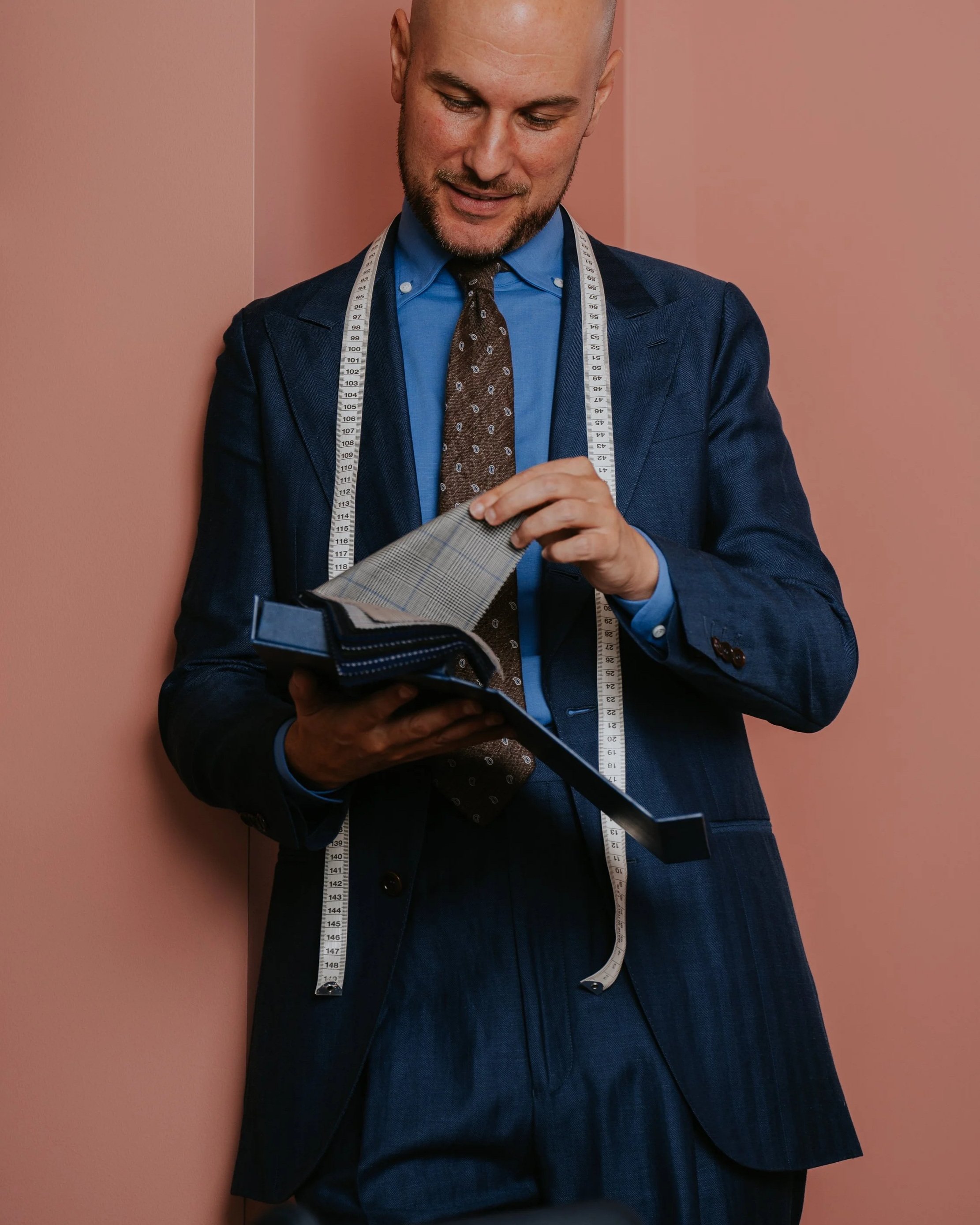 Aleksandar Stately in a Navy Herringbone made to measure suit with a measuring tape around his neck is looking and smiling at a fabric swatch in his hand.