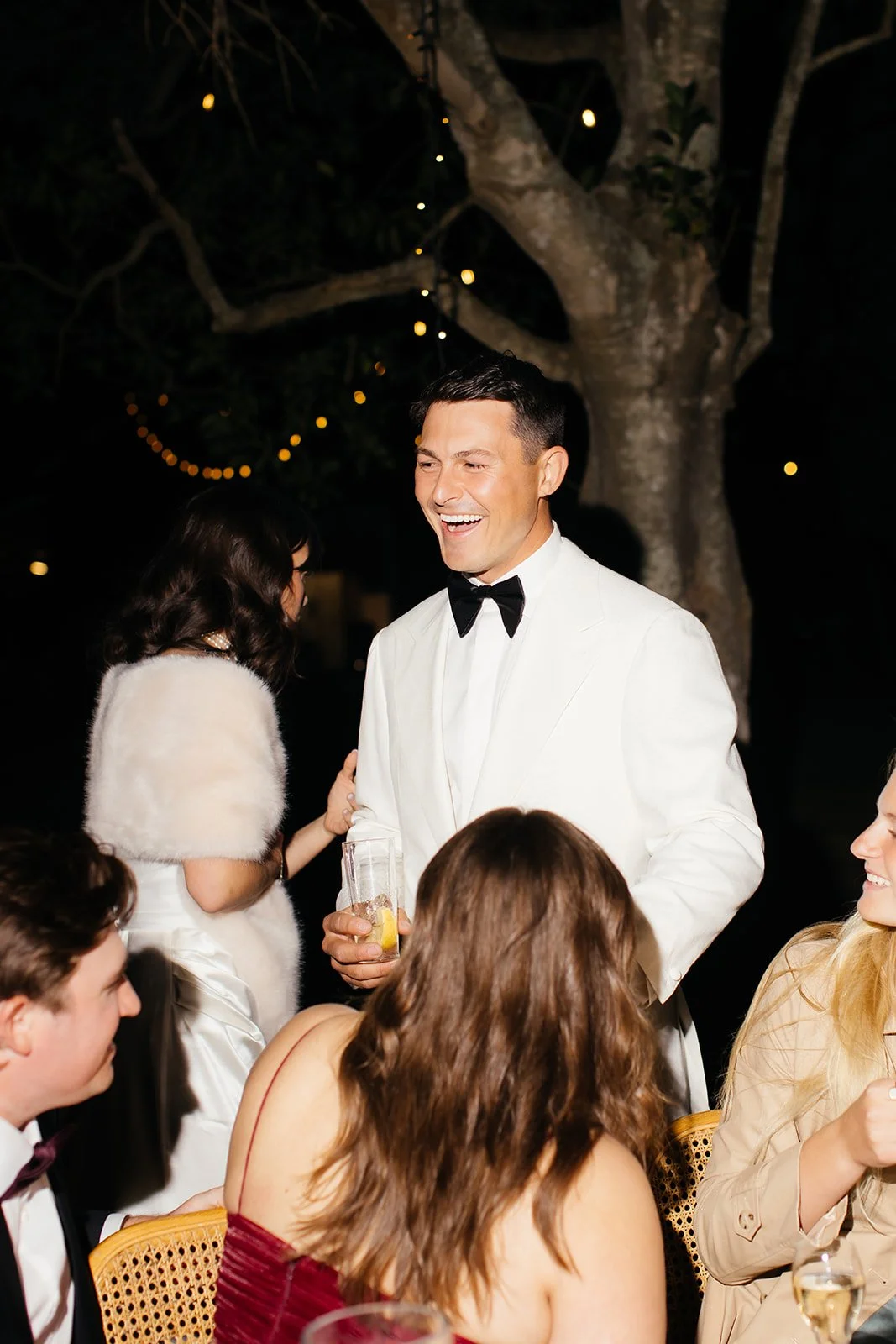 A groom in a white custom tailored made to measure Aleksandar Stately wedding tuxedo suit and black bow tie smiling and holding a glass of drinks, surrounded by women in elegant attire, under string lights and a large tree.