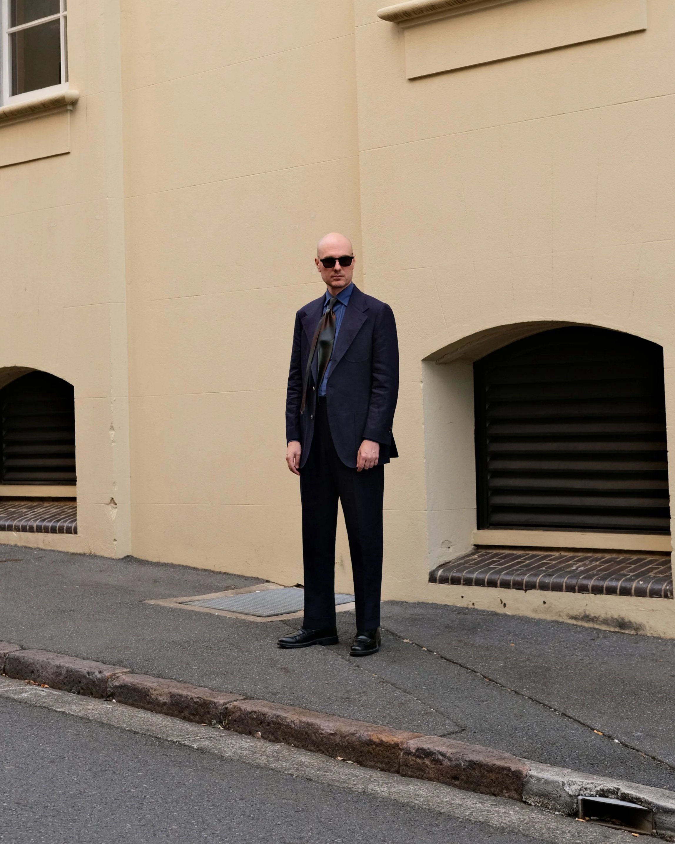 Aleksandar Stately wearing a made to measure dark navy linen suit, a blue shirt, and a tie, standing on a Brisbane city sidewalk with a beige building in the background.