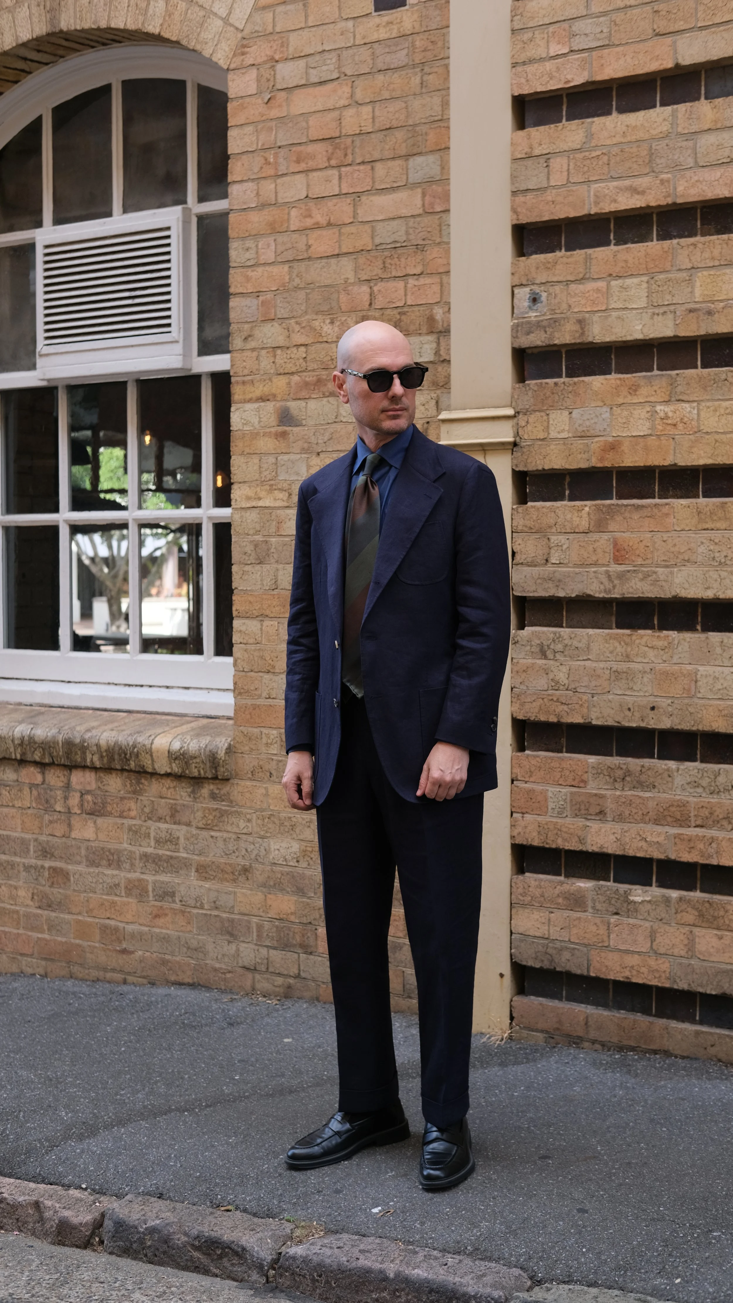 Aleksandar Stately wearing a made to measure navy linen suit, navy blue shirt, striped tie, dark sunglasses, and black loafers stands outside next to a brick wall with large windows. Brisbane Queensland