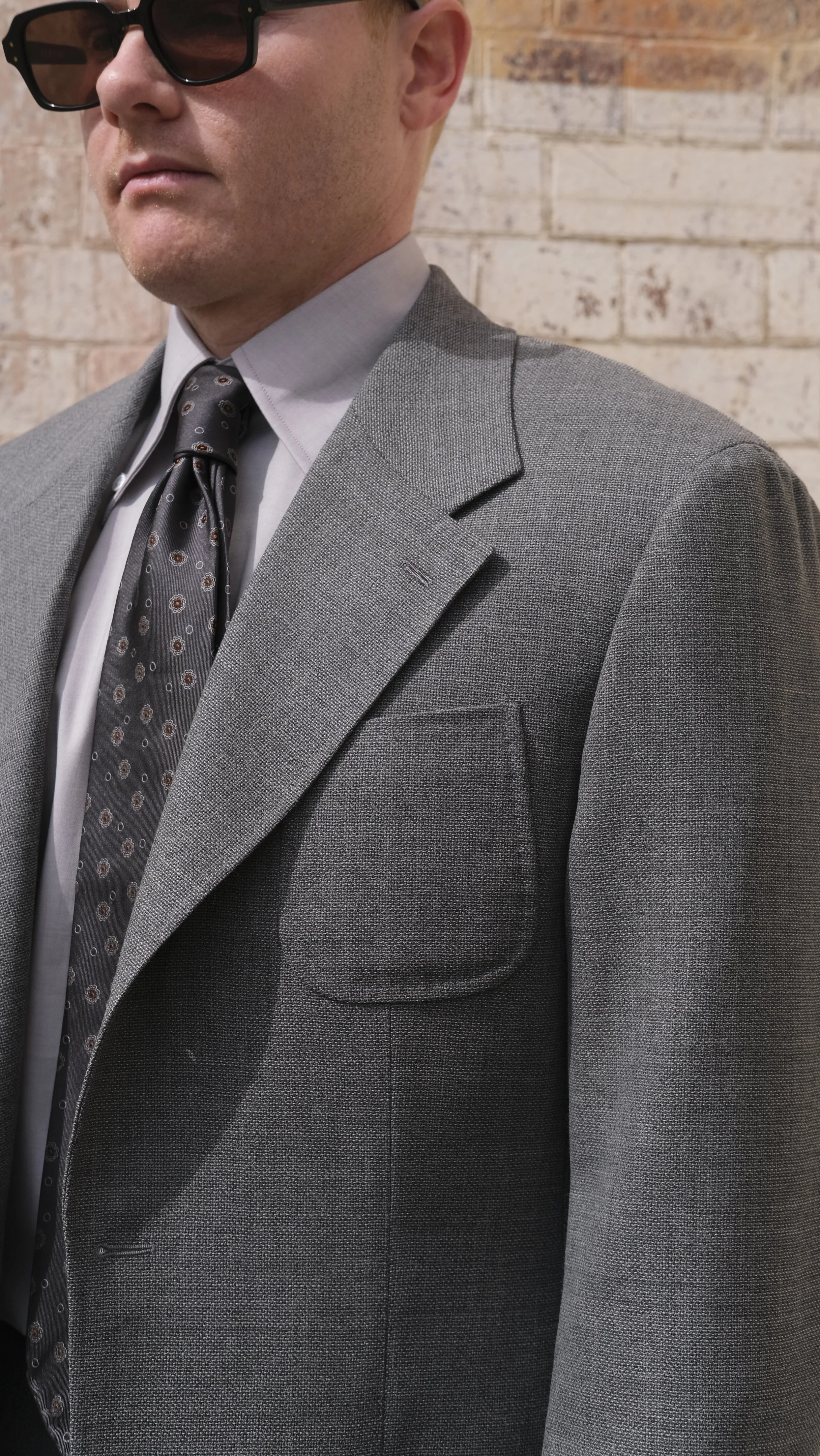 Close-up of a man wearing a custom tailor made to measure grey suit, white dress shirt, patterned tie, and black sunglasses, standing against a brick wall background.