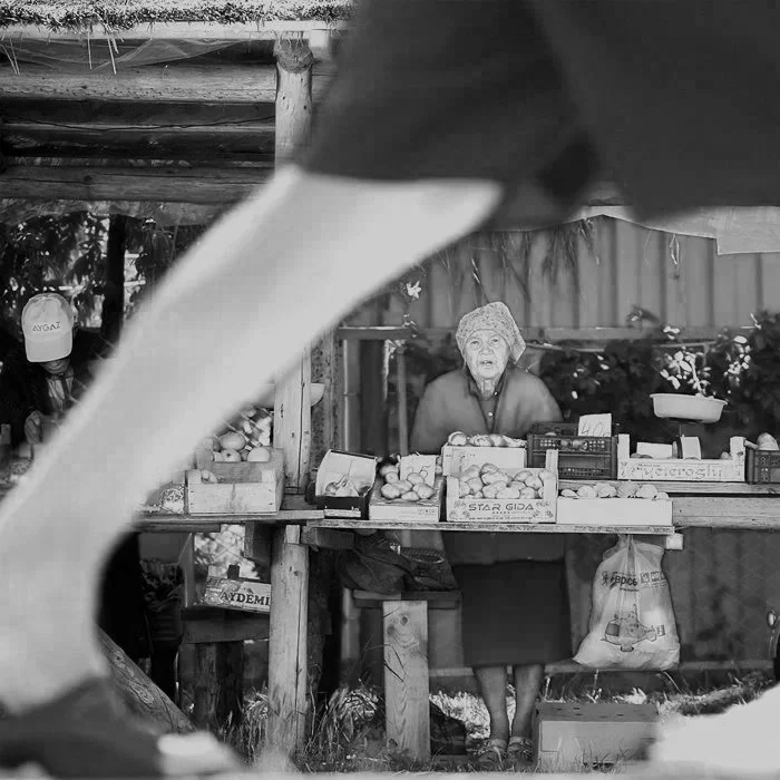 Frau steht hinter einem Obst- und Gemüsetresen in einem kleinen Marktstand, umgeben von Körben mit Früchten, im Hintergrund ein Holzzaun, Schwarz-Weiß-Foto, einige Leute im Hintergrund, eine Person trägt eine Kappe.