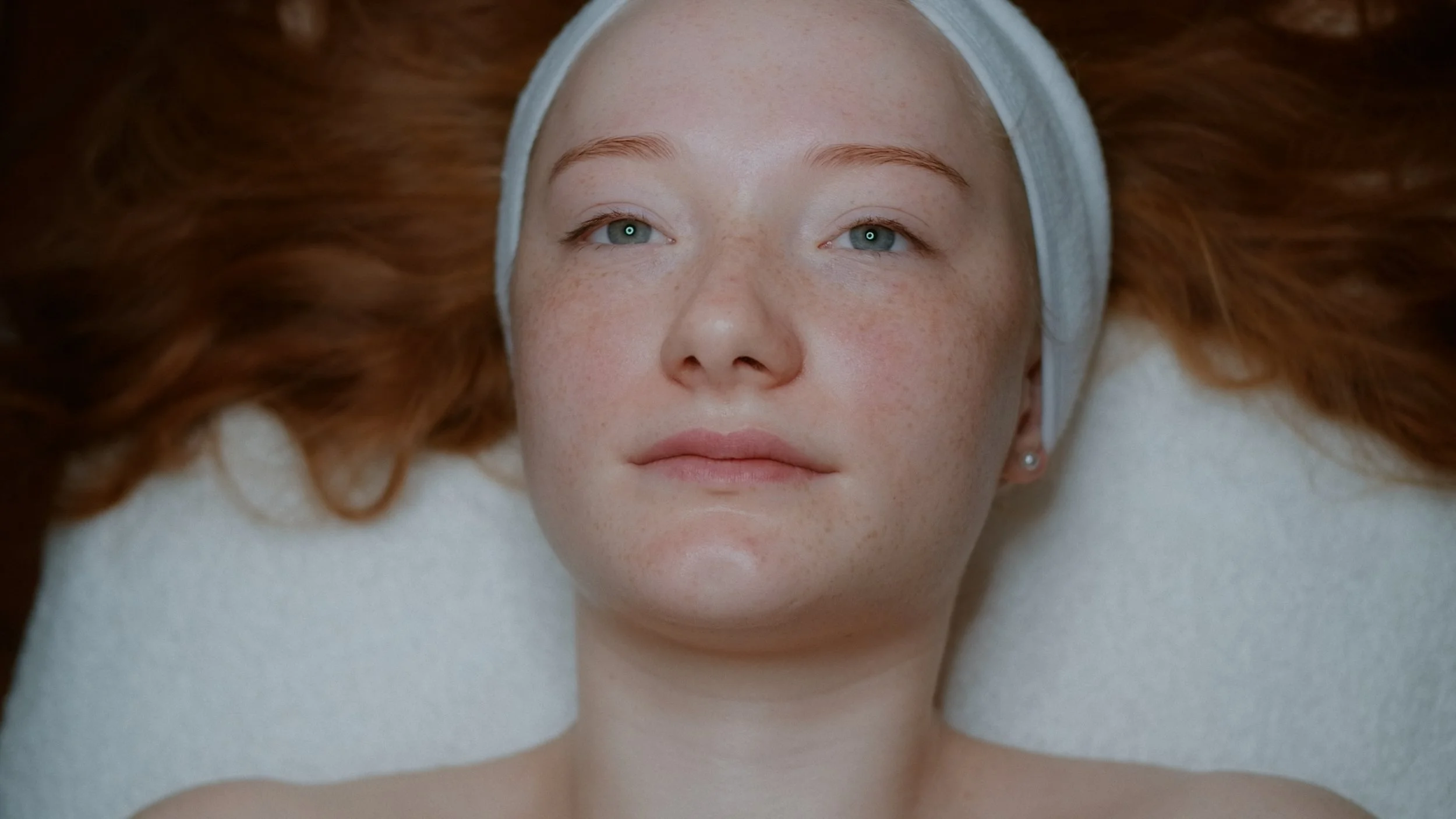 Nahaufnahme eines roten Haares, einer Frau mit blauen Augen, hellen Sommersprossen und entspanntem Gesichtsausdruck, liegend auf einem weißen Untergrund mit einem weißen Stirnband.