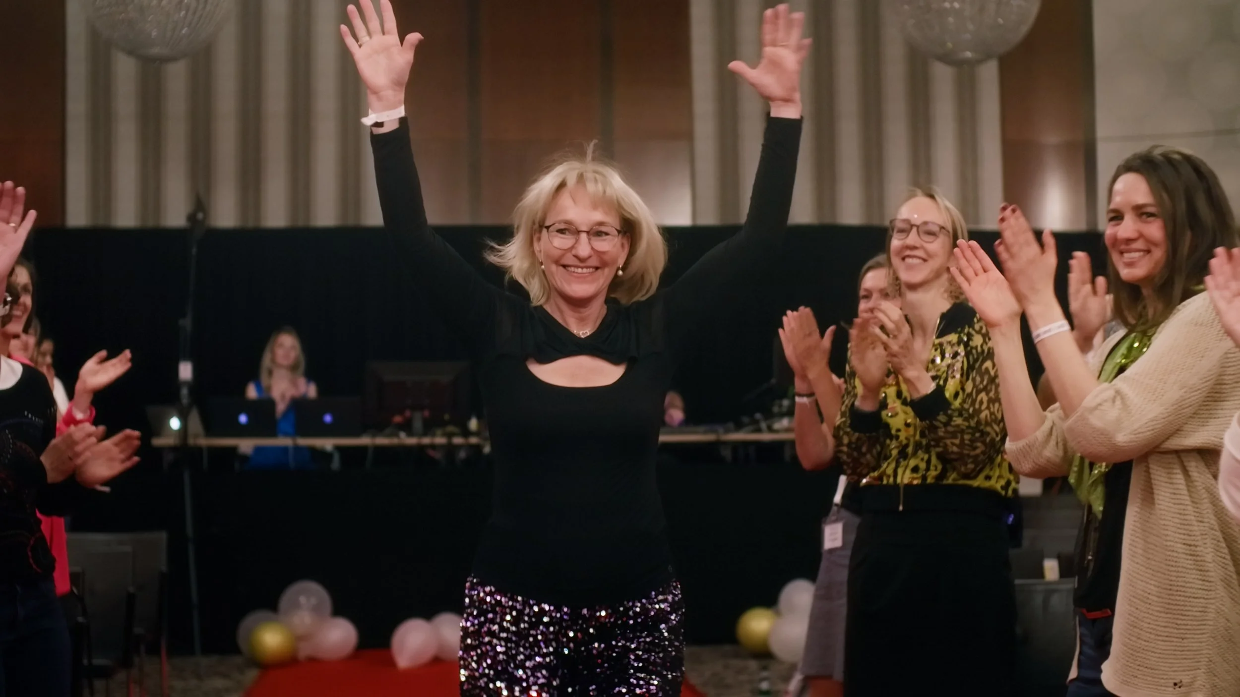 Eine lachende Frau in einem schwarzen Kleid mit Glitzerrock steht mit erhobenen Armen auf einer Feier. Sie ist umgeben von applaudierenden Frauen, die sich freuen. Im Hintergrund sind Tanzflächen, Dekorationen und DJ-Equipment zu sehen.