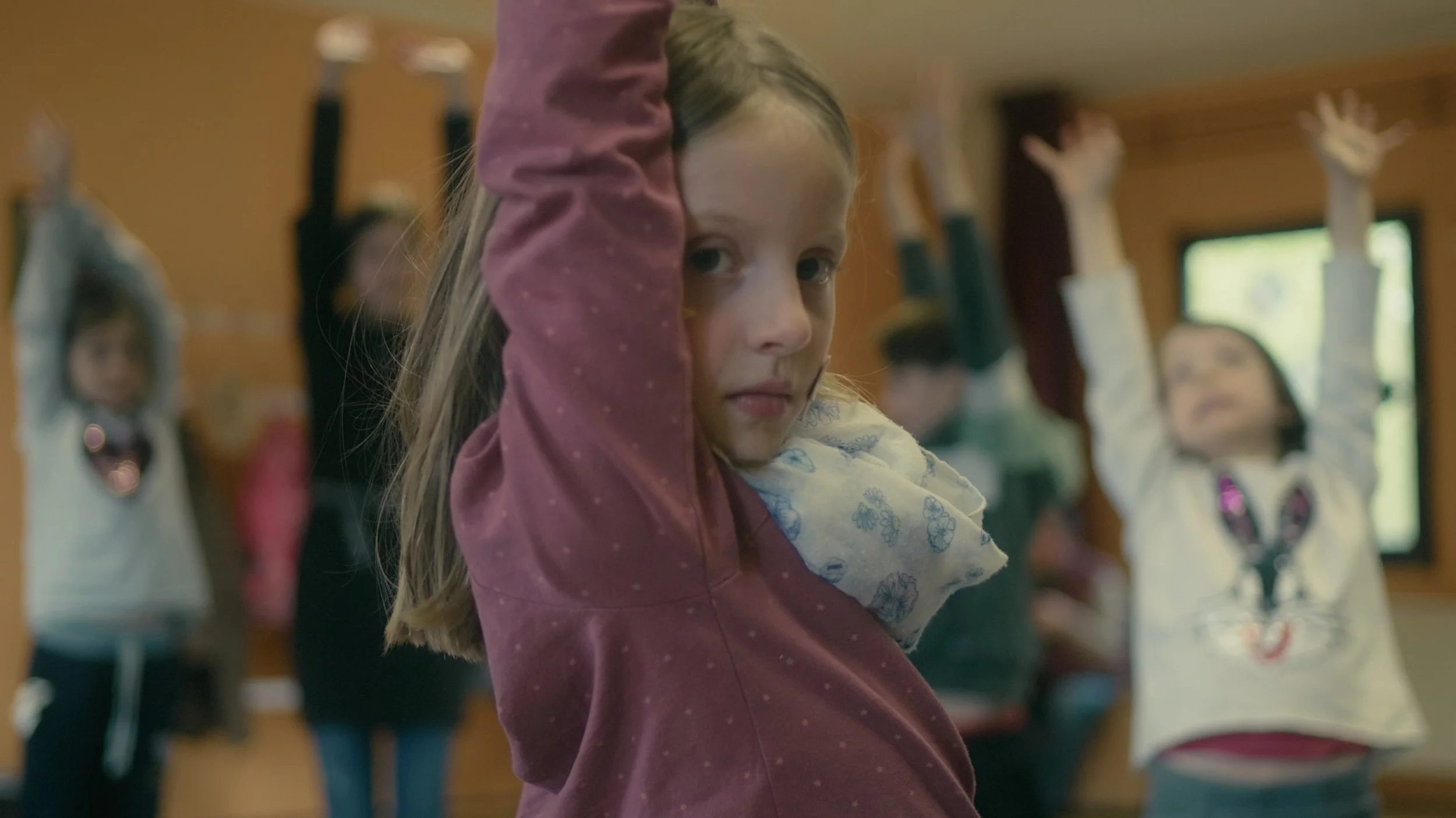 Ein junges Mädchen mit langem, blondem Haar, das in einem Klassenzimmer nach vorne schaut, während andere Kinder im Hintergrund den Arm heben.