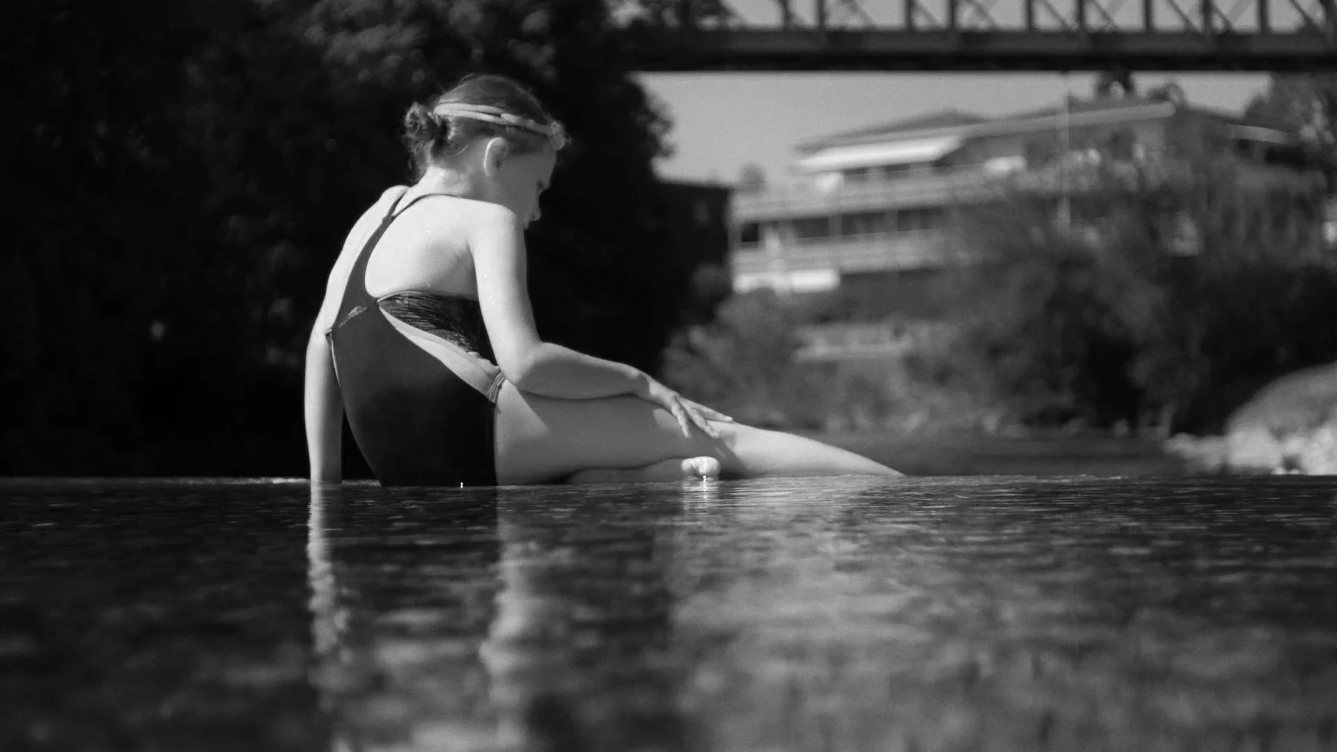 Ein junges Mädchen in einem Badeanzug sitzt an einem Wasserufer, mit Blick auf das Wasser, im Hintergrund sind Häuser und Bäume sichtbar.