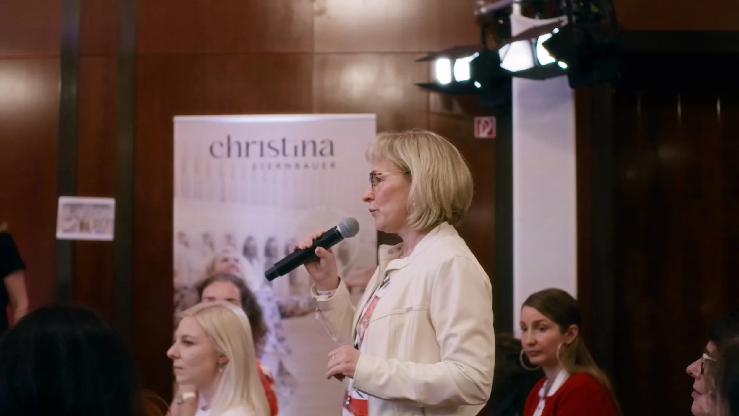 Eine Frau mit Brille spricht in ein Handmikrofon während einer Veranstaltung. Im Hintergrund steht ein Banner mit dem Namen Christina Bukerbauer.