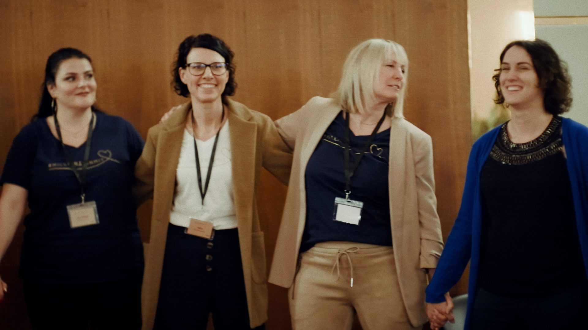 Vier Frauen stehen nebeneinander und halten sich an den Händen, lachen und wirken fröhlich bei einer Veranstaltung.