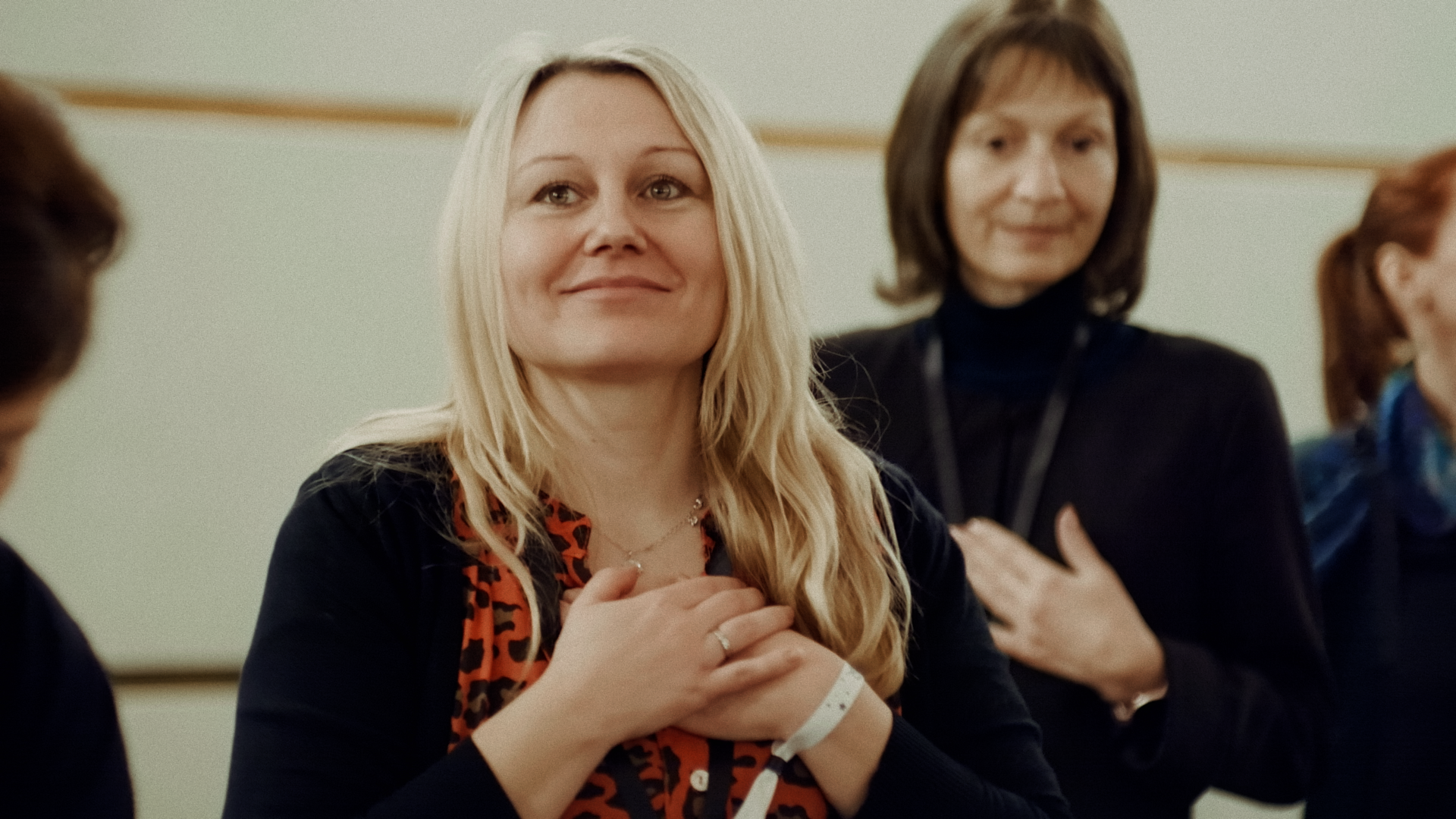 Drei Frauen sitzen in einem Raum und scheinen aufmerksam zuzuhören oder zu sprechen, die mittlere Frau lächelt sanft und hat die Hände auf der Brust. Die Frauen tragen dunkle Kleidung, und im Hintergrund ist eine helle Wand.
