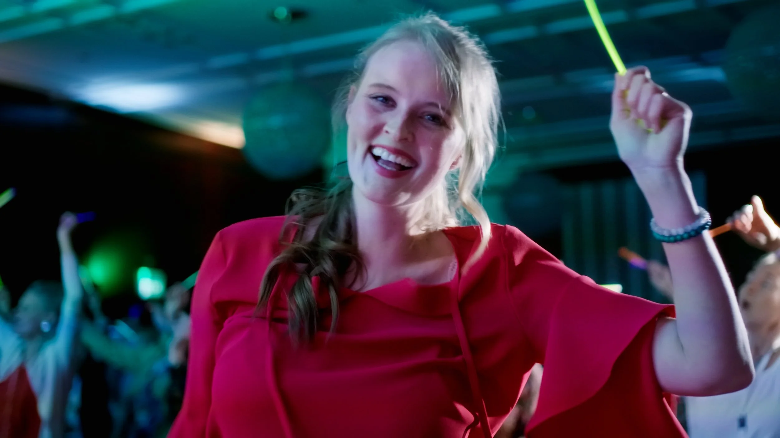 Junge Frau tanzt und lacht in einer Disco mit bunten Lichtern, trägt einen roten Pullover.