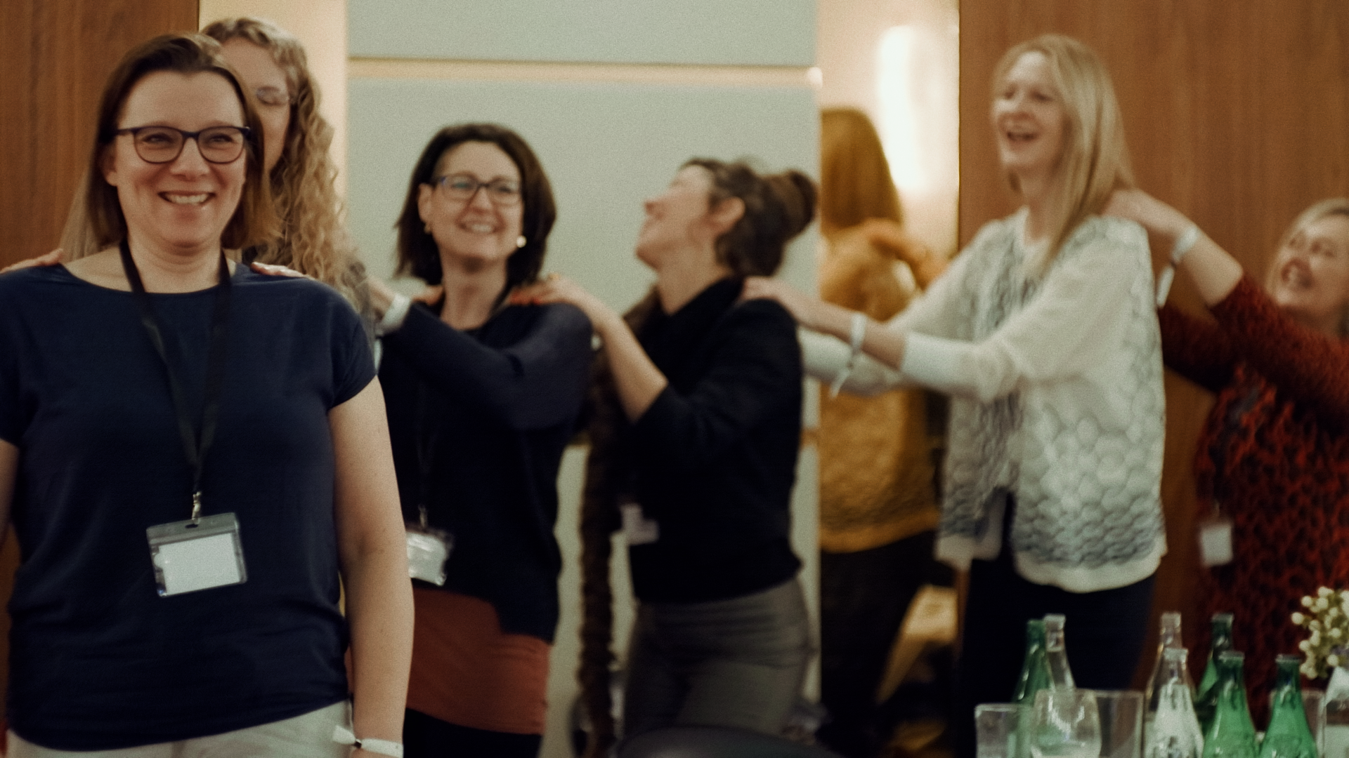 Gruppen von lachenden Frauen bei einer Teambuilding-Aktivität, bei der sie sich in einer Kette verbinden, im Innenraum einer Veranstaltungshalle.