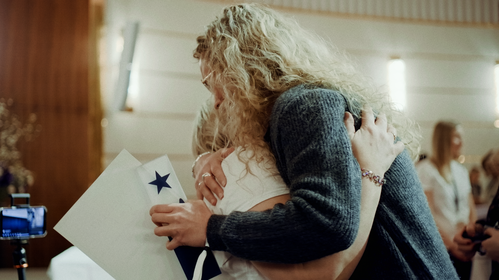 Zwei Frauen umarmen sich emotional bei einem feierlichen Anlass, eine hält ein Geschenk, im Hintergrund weitere Personen.