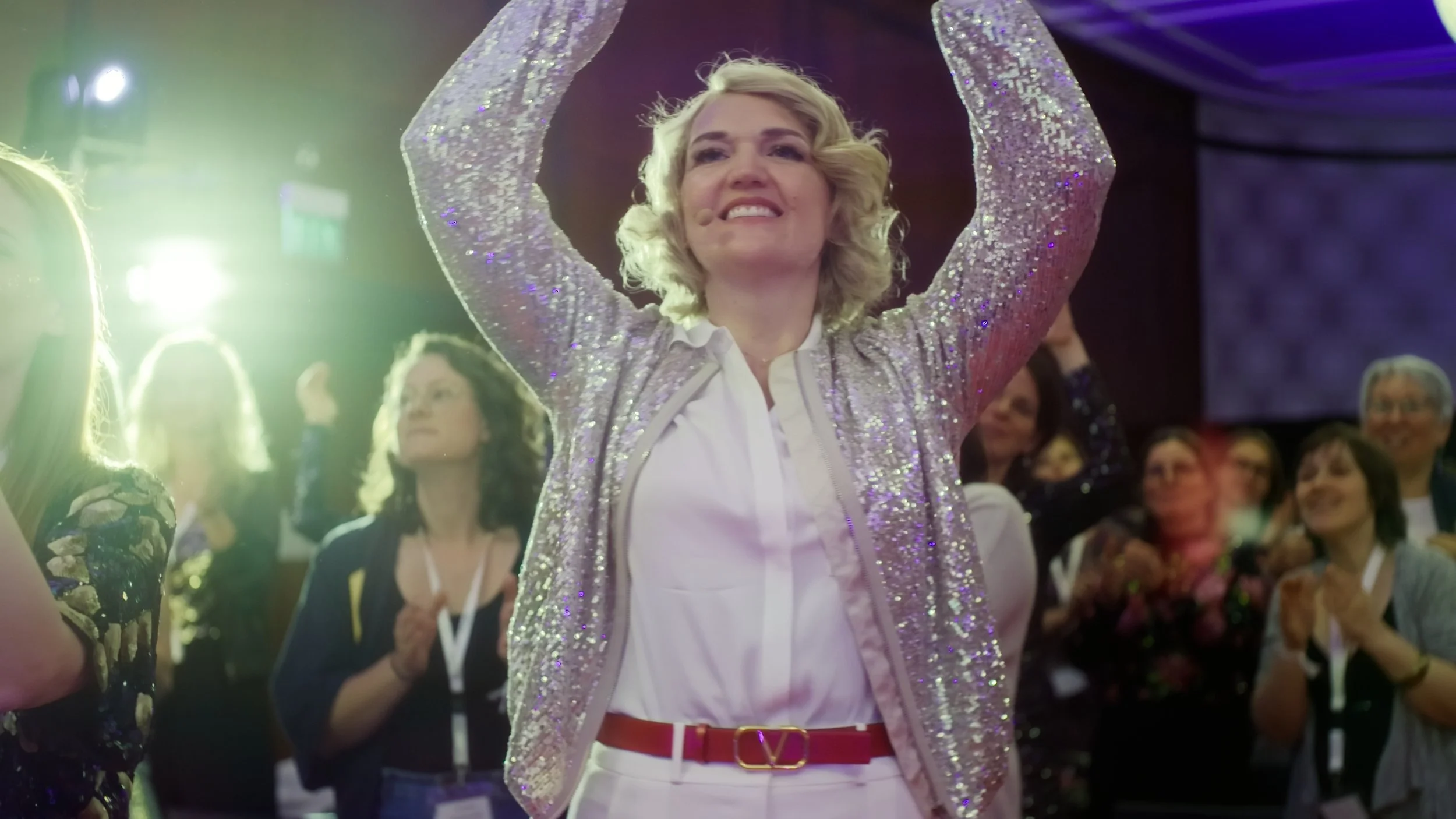 Eine lachende Frau mit blonden Locken trägt ein glitzerndes, perlenbesetztes Oberteil und zeigt Felizität auf einem Konzert oder Event. Im Hintergrund sind mehrere stehende Menschen, die ebenfalls applaudieren und das Ereignis beobachten.