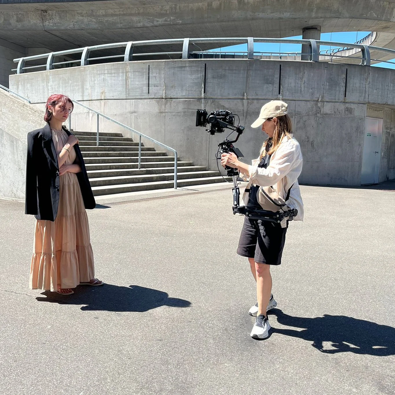 Eine Frau filmt eine andere Frau mit einer professionellen Kamera auf einer Straße im Freien, vor einer betonnenen Treppe und einer modernen Brücke, bei sonnigem Himmel.