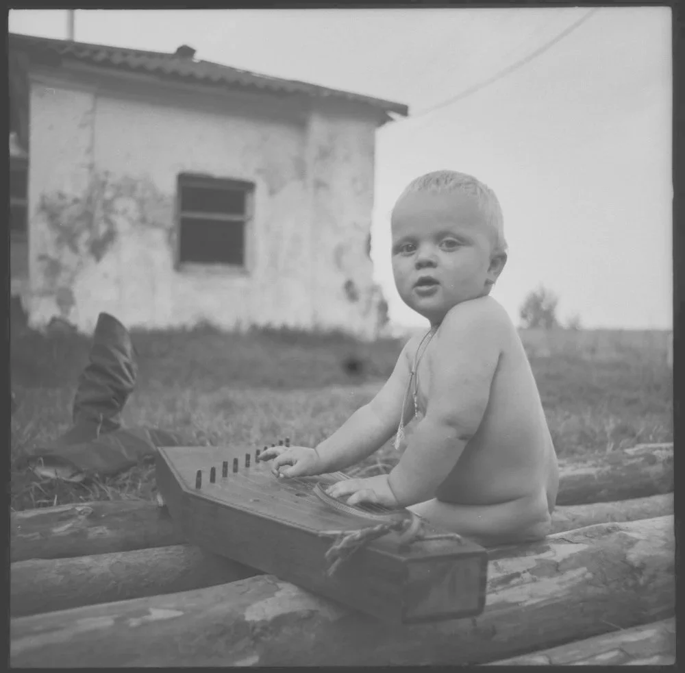 Ein nacktes Baby, das auf Holzbalken sitzt und eine kleine Wurlitzer-Orgel spielt, im Freien vor einem alten verfallenen Haus.