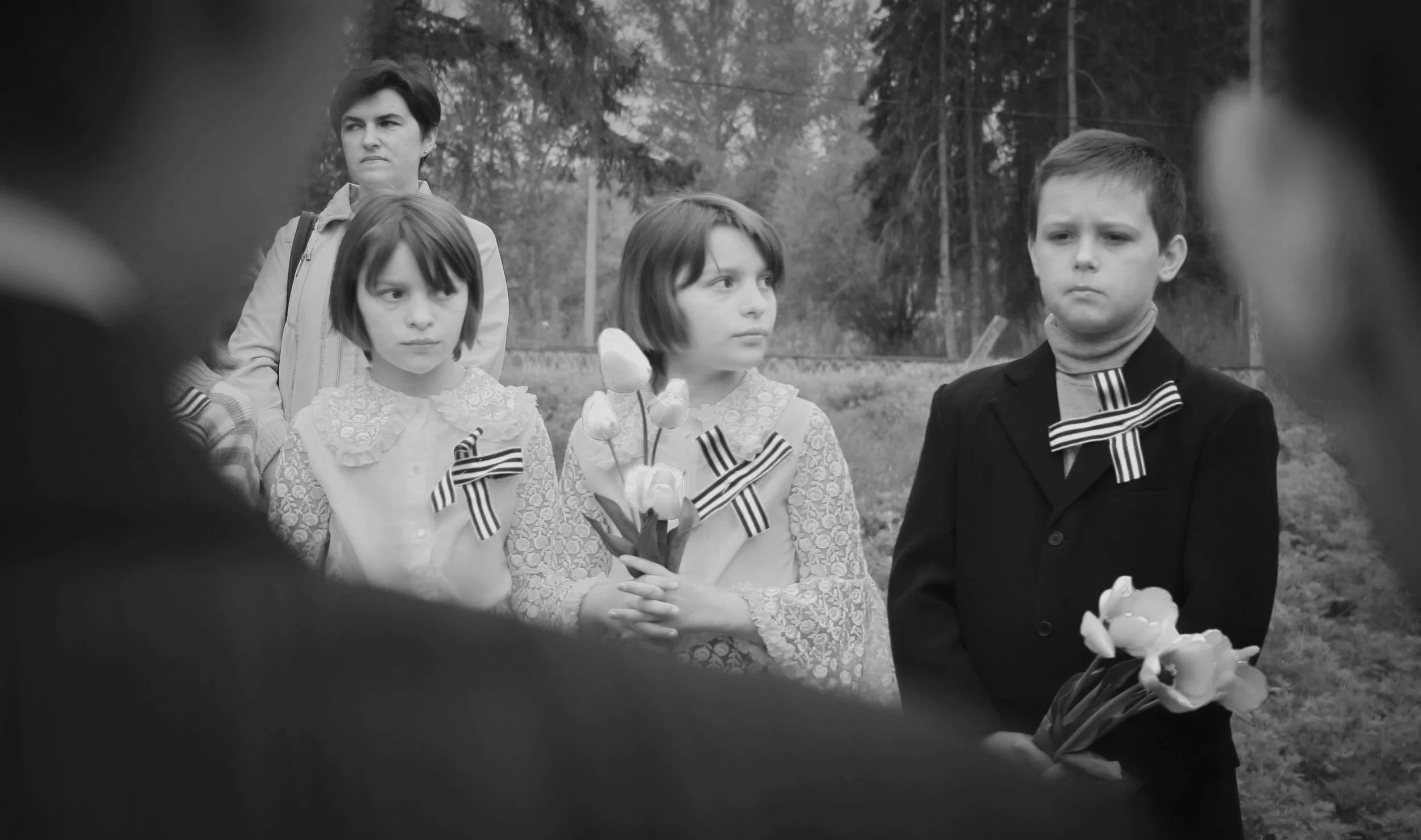 Schwarz-weiß-Foto von vier Kindern, die an einem Tag im Freien stehen, zwei Mädchen mit Blumenmusterkleidern und Knopfleiste, ein Junge in Anzug mit Fliege sowie ein Erwachsener im Hintergrund, alle mit gestickten schwarzen und weißen Schleifen am Kleidungsstück. Die Szene wirkt ernst, und es scheint eine formelle Veranstaltung oder eine Gedenkfeier zu sein.