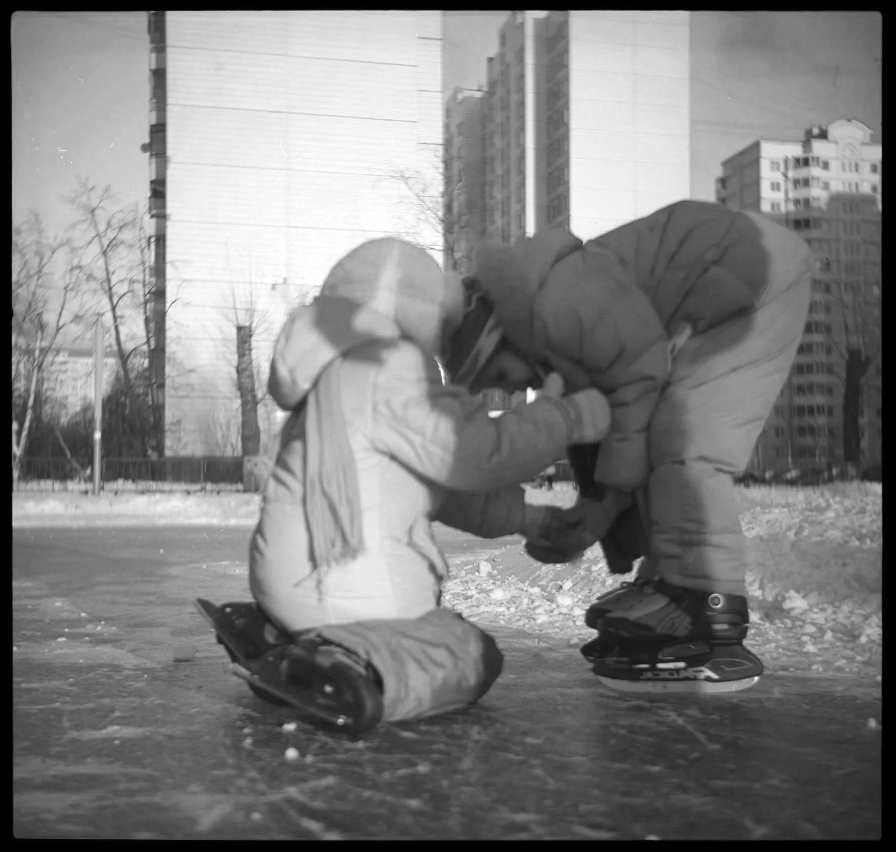 Ein Erwachsener hilft einem Kind beim Eiskunstlaufen im Freien auf einer eisbedeckten Fläche, mit Gebäuden im Hintergrund.