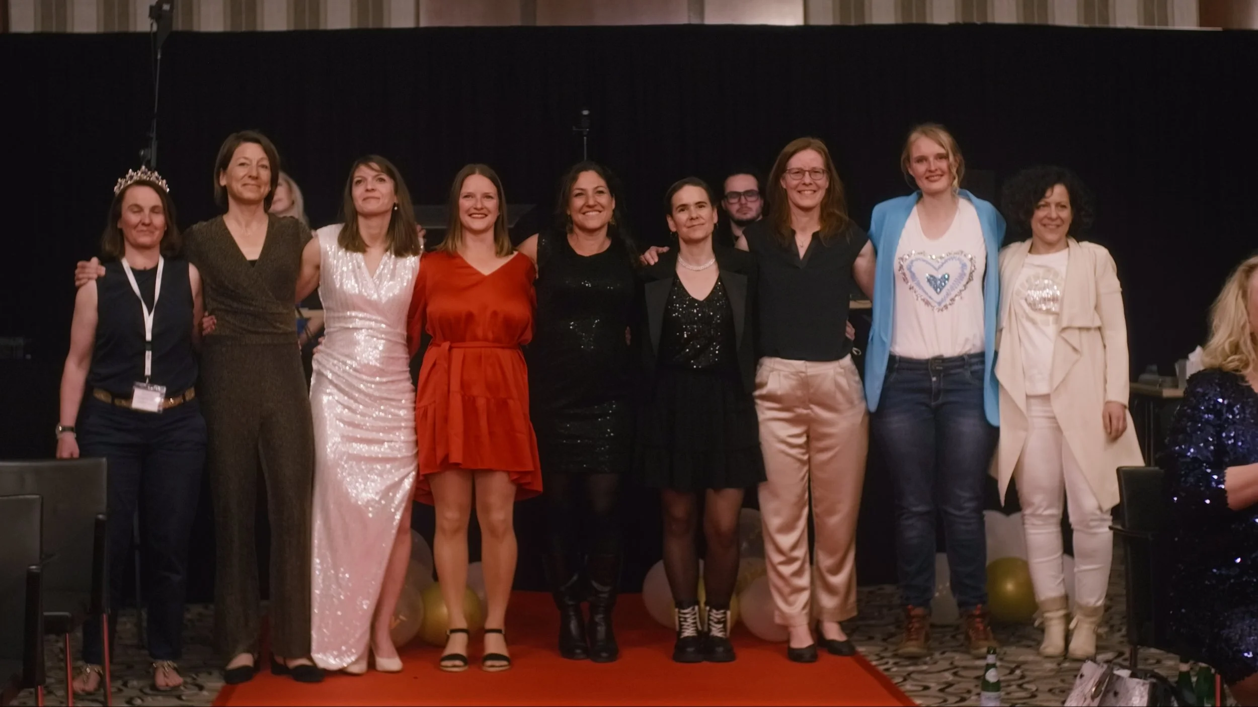 Gruppe von Frauen und Männern in festlicher Kleidung auf einer Bühne, einige lachen, einer trägt eine Krone.