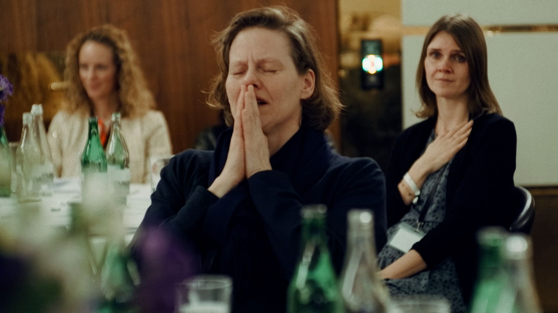 Drei Frauen sitzen an einem Tisch bei einer Veranstaltung. Die Frau in der Mitte hat die Augen geschlossen und die Hände betend, während die Frauen im Hintergrund die Hand auf die Brust gelegt haben.