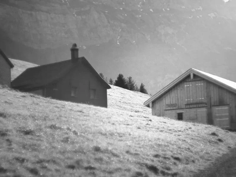 Schwarz-weiß-Bild von Hütten auf einem schneebedeckten Hang, mit Bäumen im Hintergrund.
