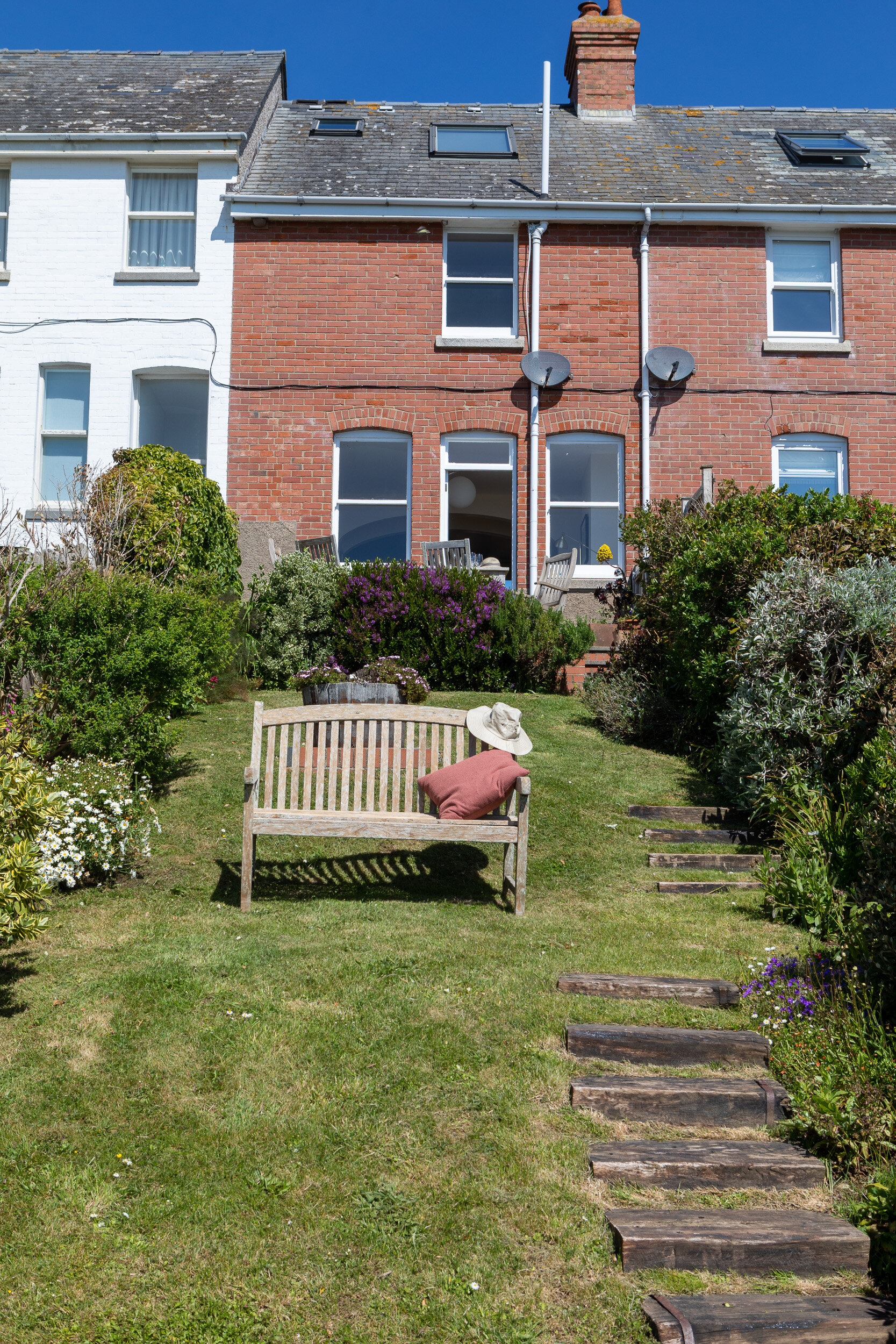 Backyard garden with wooden bench, hat, and cushion facing a brick and white terraced house, with a stone path and greenery.