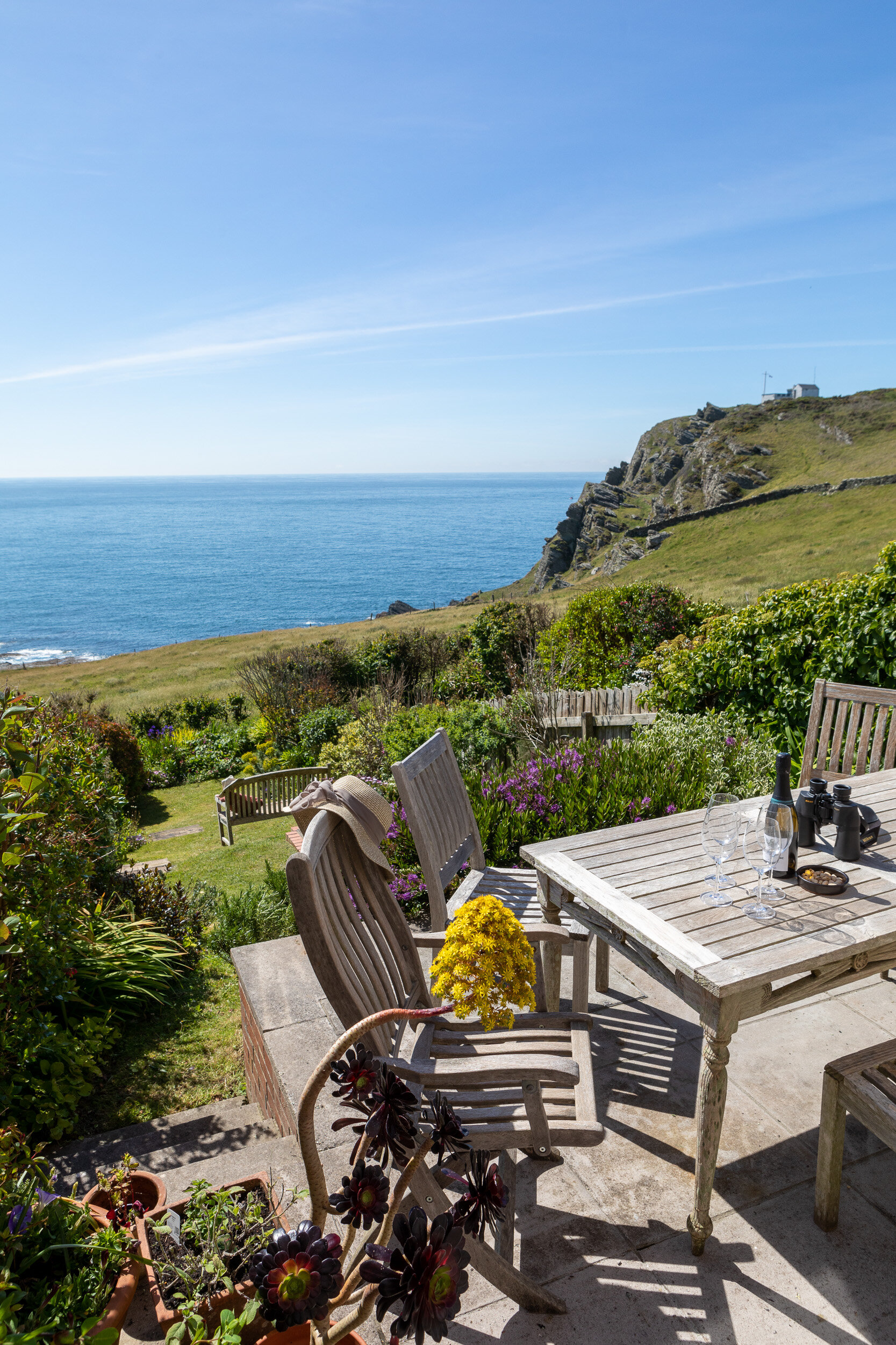 Outdoor seating area with a wooden table and chairs overlooking a scenic coastal view; small garden with flowers and potted plants.