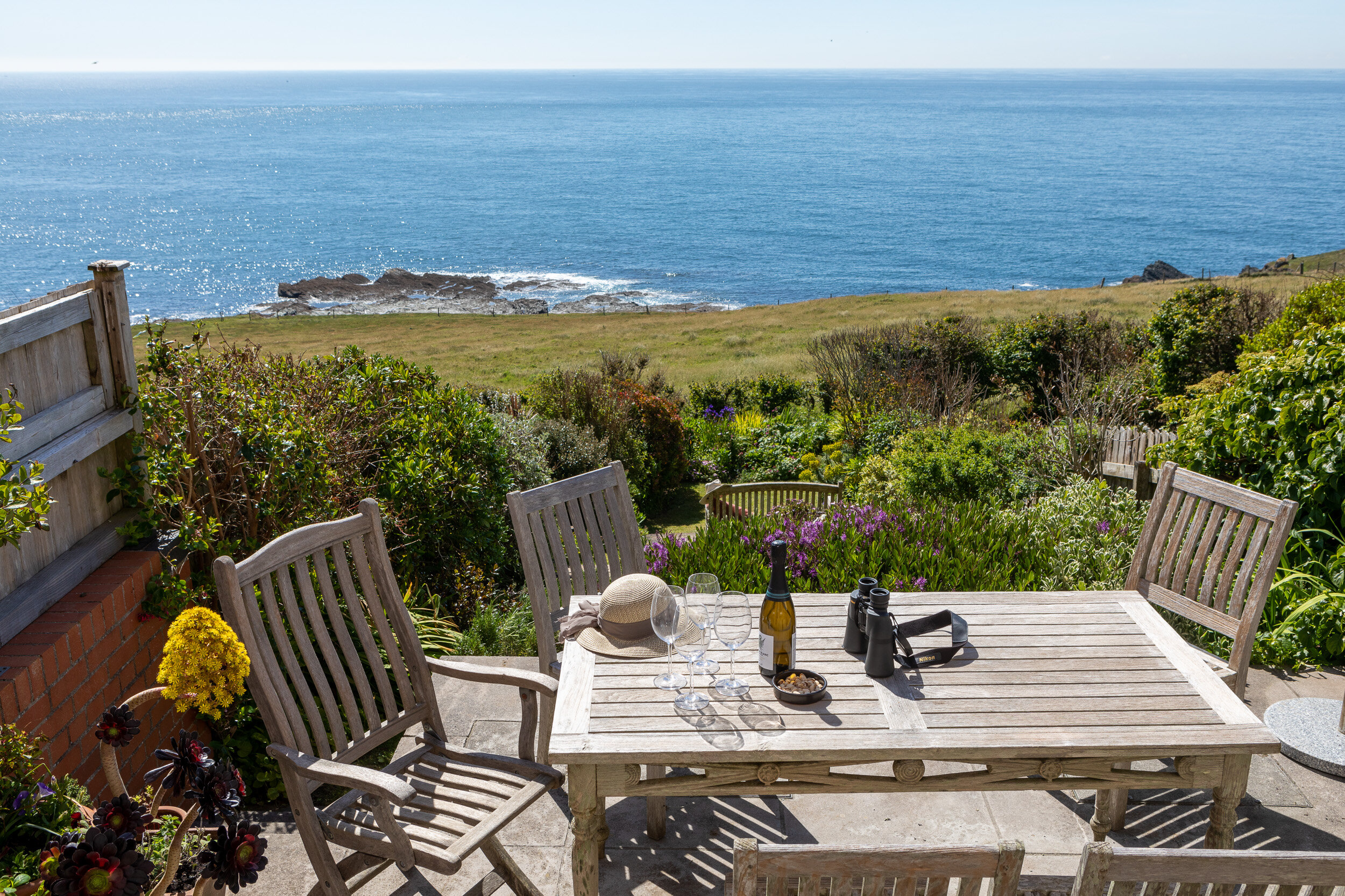 Outdoor wooden table and chairs set in garden overlooking ocean, with wine bottle, glasses, binoculars, and hat on table.