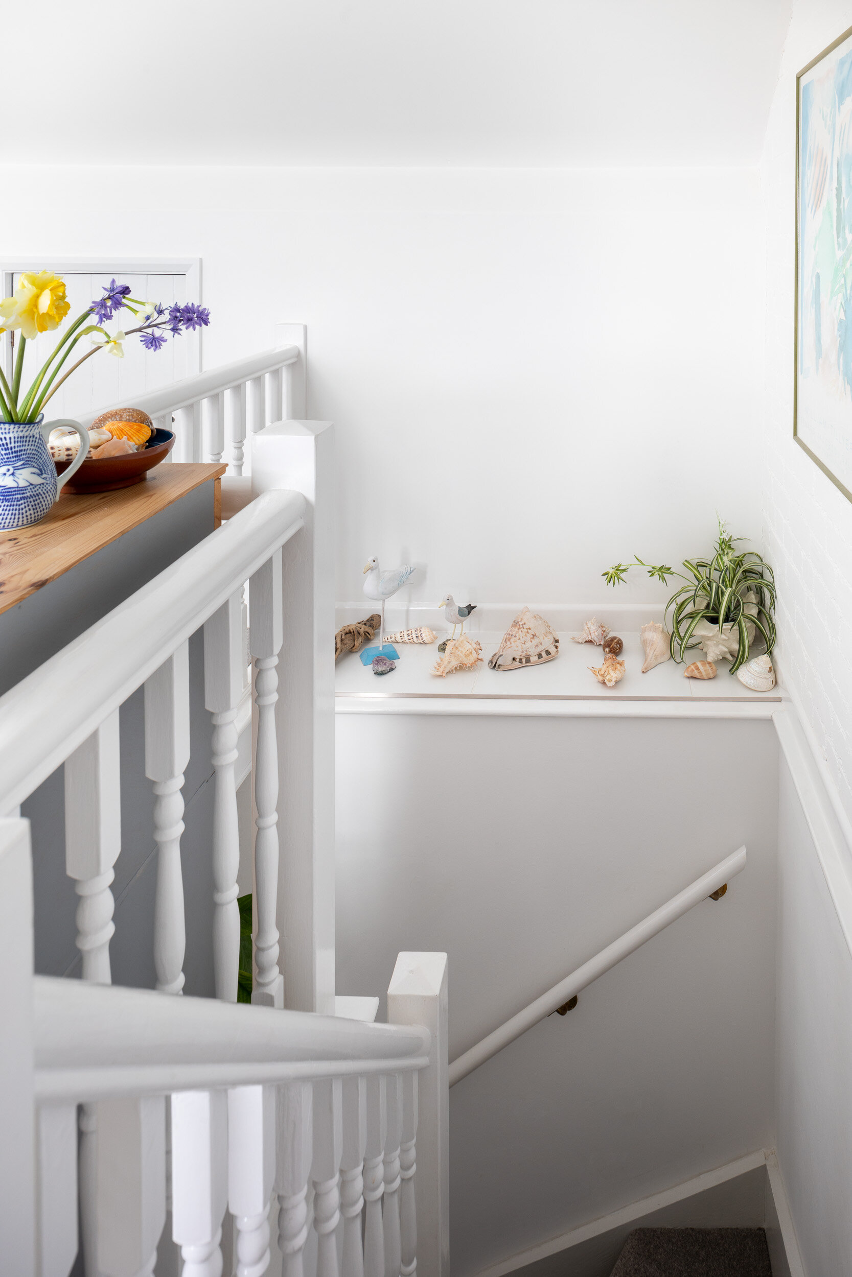 Interior of a white staircase with decorative items on the ledge including sea shells, bird sculptures, and a plant, with a vase of flowers and a fruit bowl on a nearby side table.