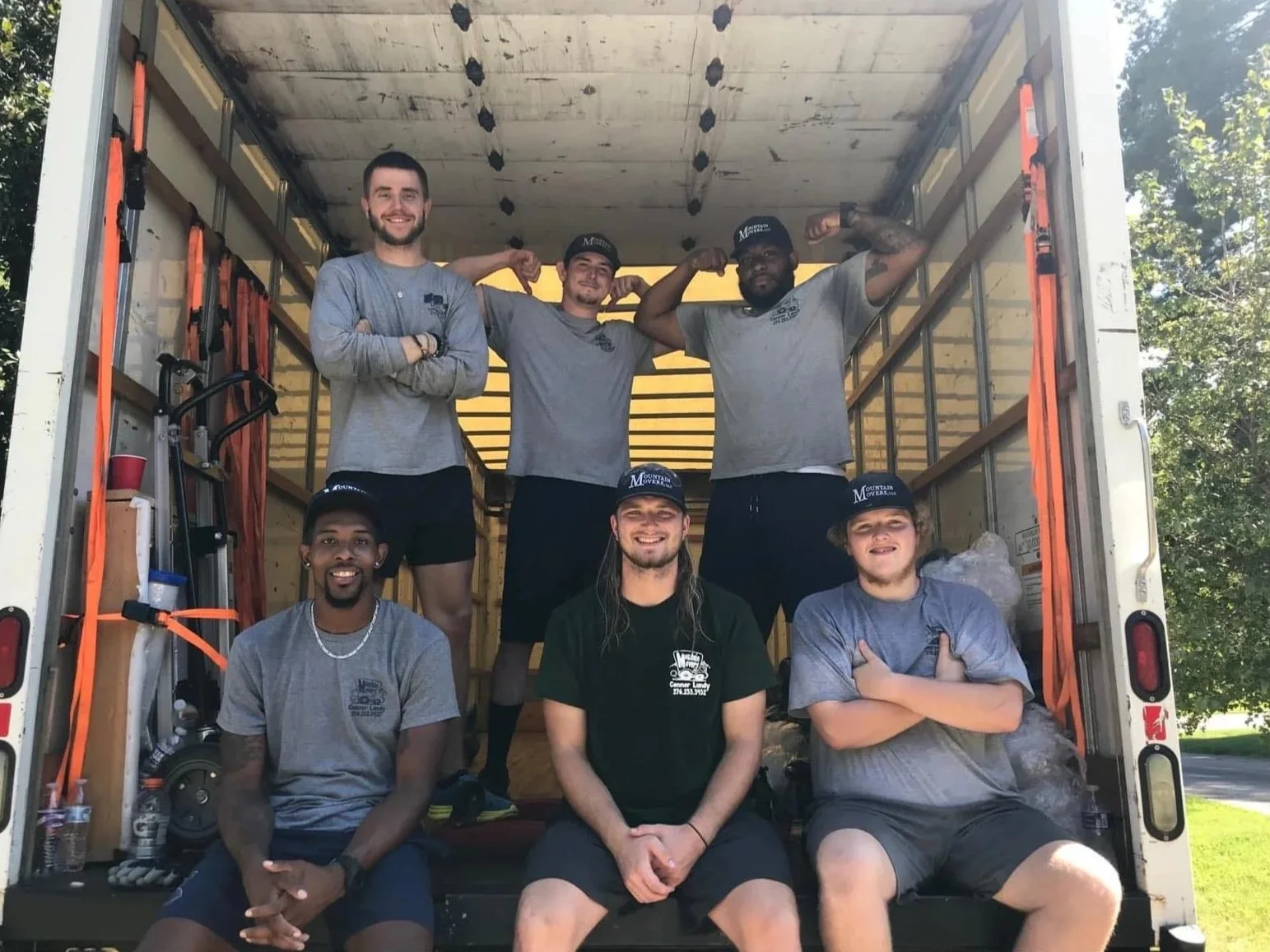 Group of six people in a moving truck, some sitting and some standing, smiling and showing muscles, wearing casual clothes and caps that say 'Mountain Movers.'