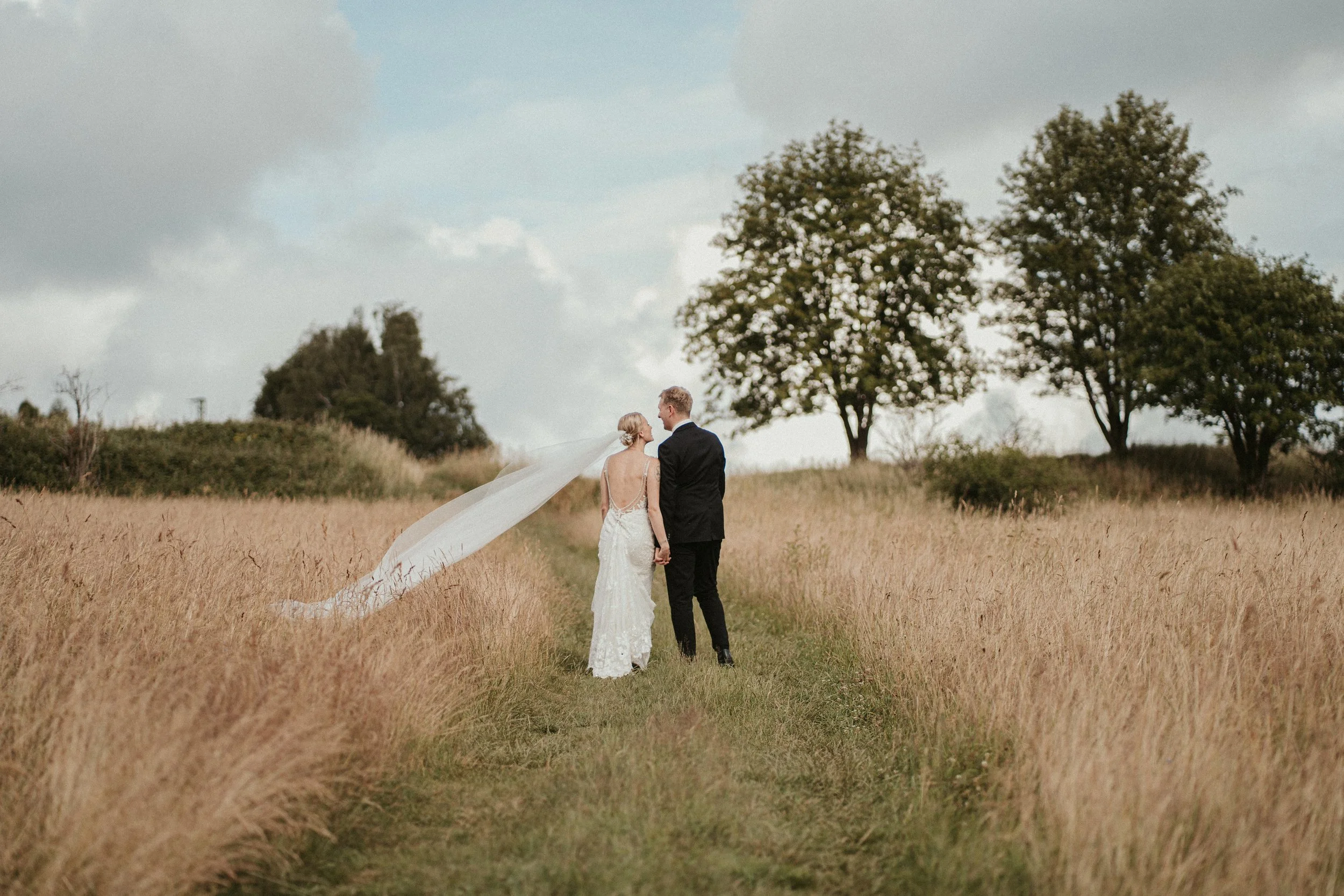 A bride and groom holding hands in a field with tall grass, with trees and cloudy sky in the background.