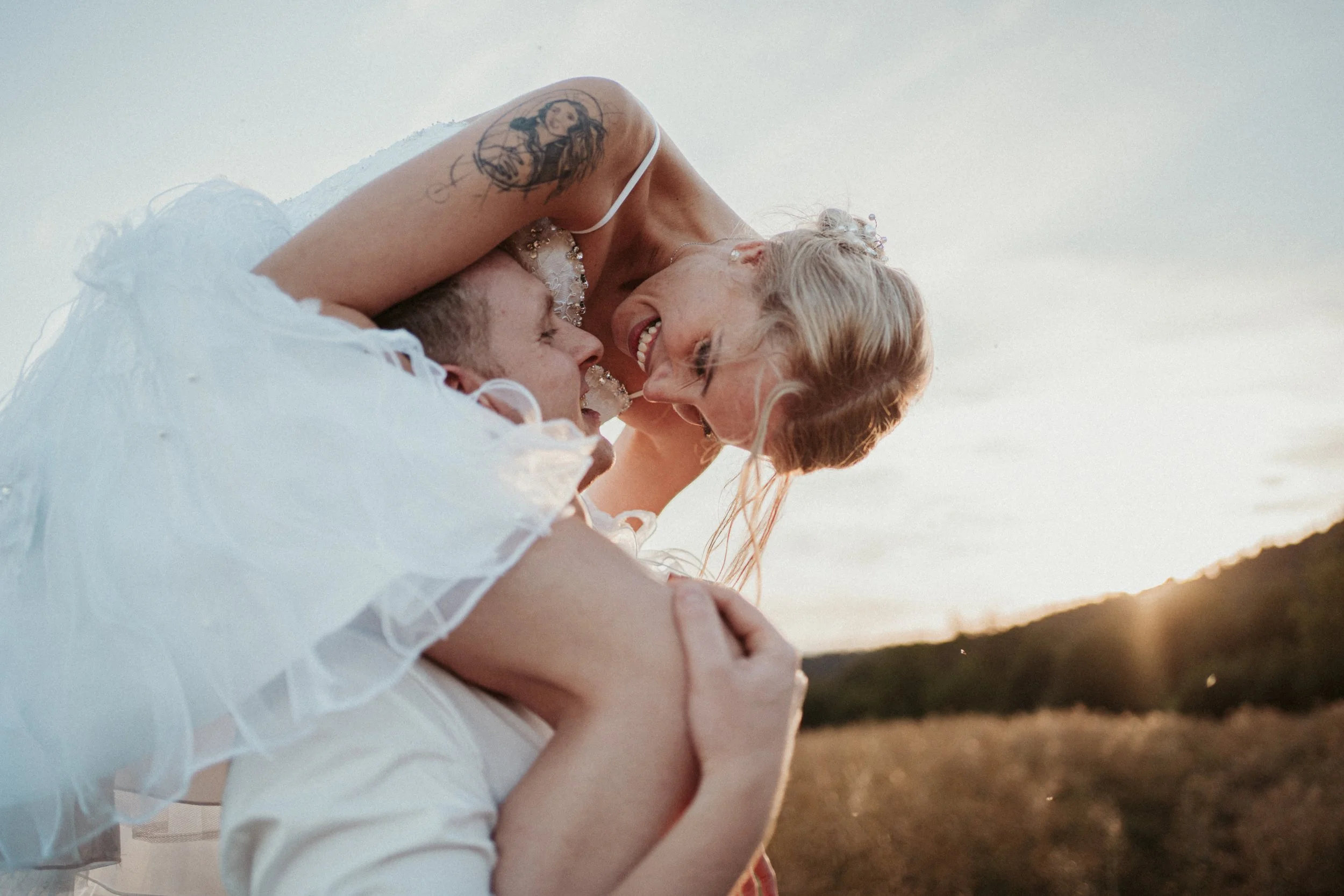 A woman with short blonde hair and tattoos on her arm is lying on the back of a man, talking and smiling in an outdoor setting during sunset.
