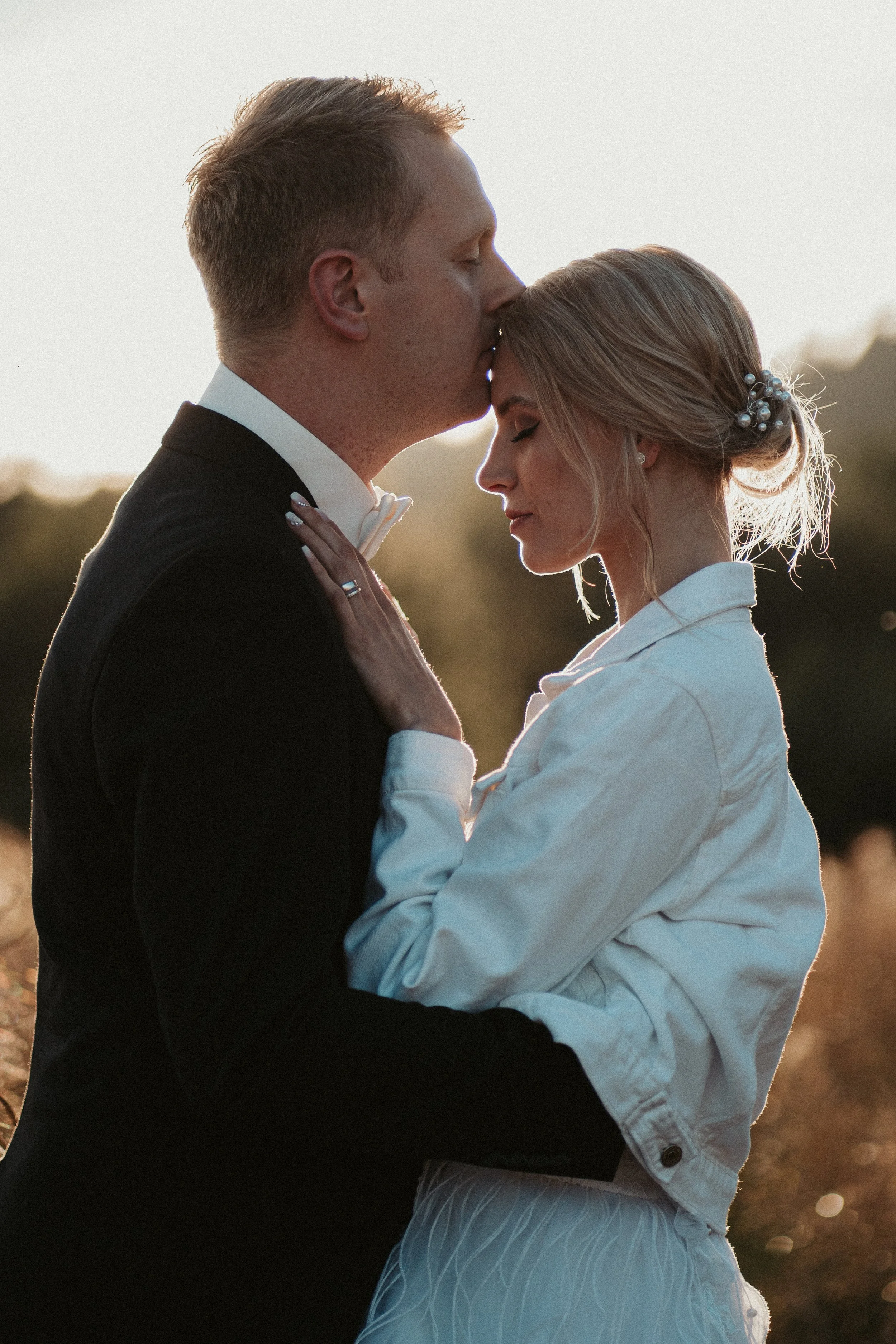 A couple in formal attire, with the man kissing the woman on the forehead, during sunset outdoors.
