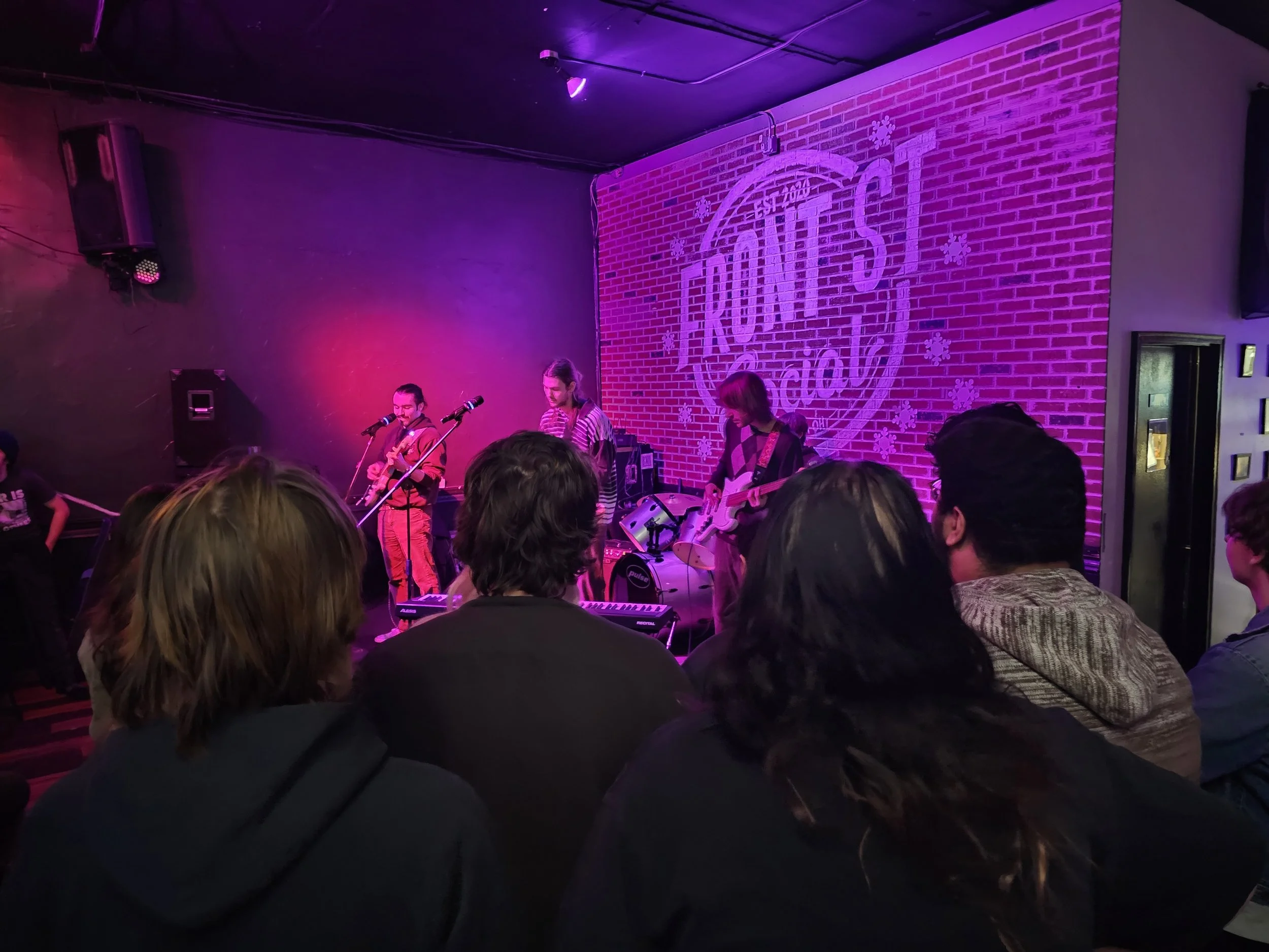 Live band performing under purple lights at a crowded indoor concert venue.