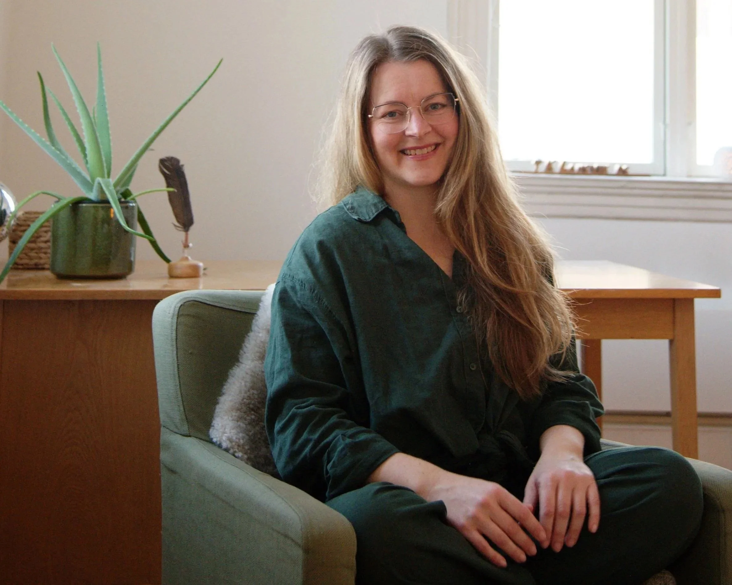 Et bilde av en smilende kvinne med langt, lyst hår og briller, som sitter i en lenestol. Hun heter Tone Langvik, er kraniosakralterapeut og innehaver av VÅGE. Bak henne er en aloe vera-plante på et bord, og et vindu.