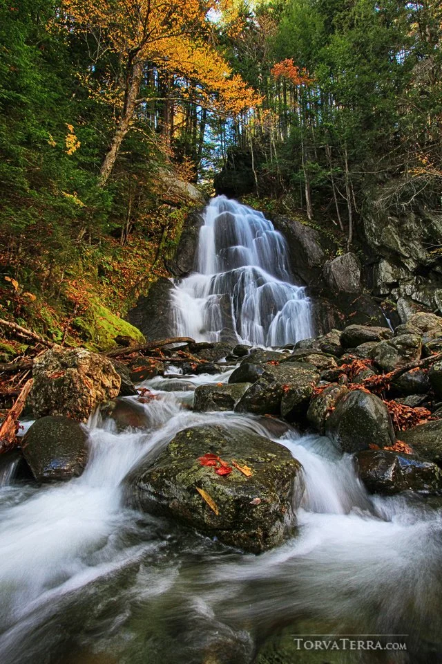A waterfall flowing over rocks surrounded by autumn trees with colorful leaves.