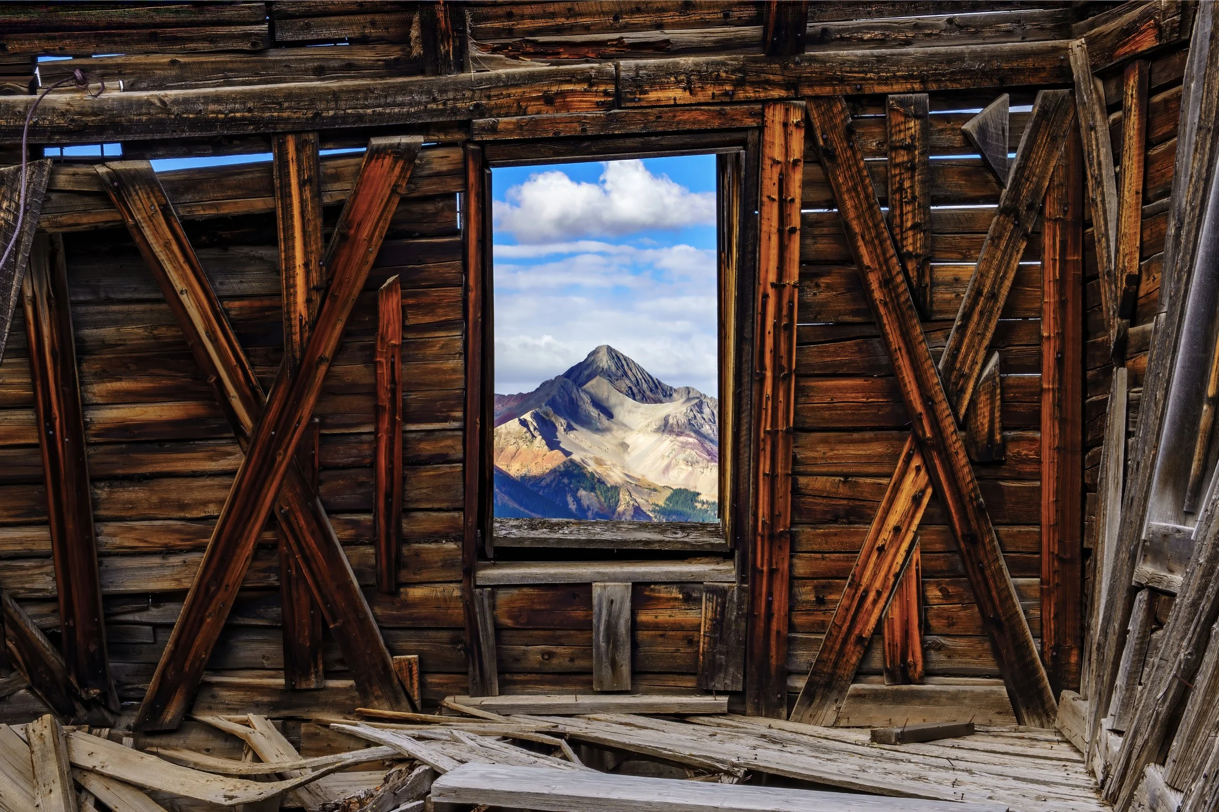 View of a mountain through the window of a dilapidated wooden building with broken walls and fallen debris on the floor.