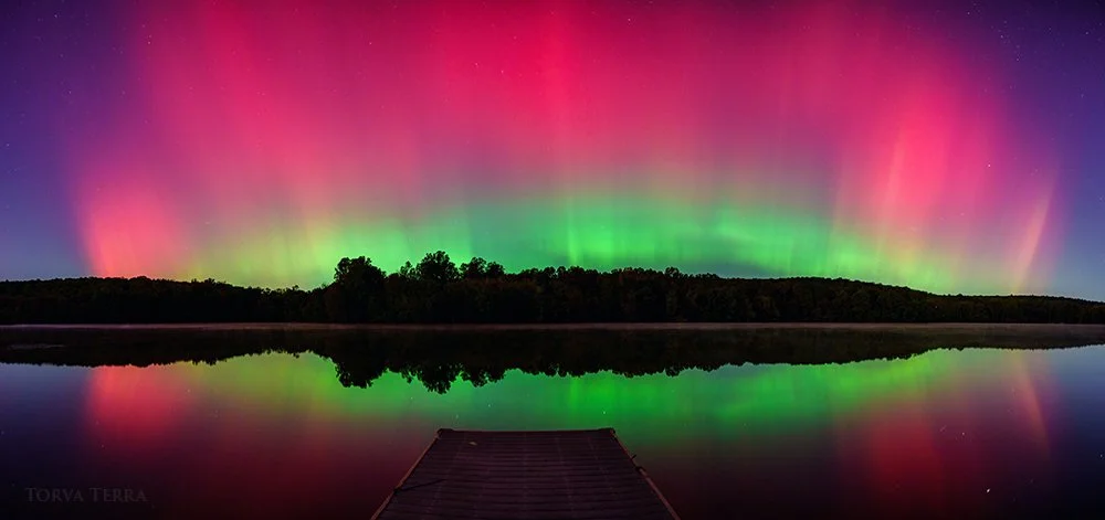 Aurora borealis above a lake reflecting colorful lights with a wooden dock in the foreground.