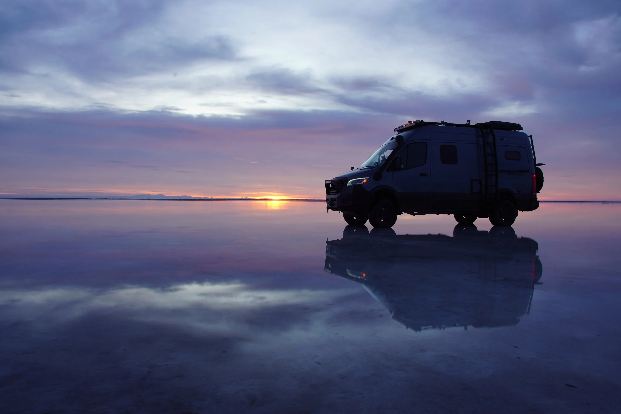 A black camper van parked on a reflective salt flat at sunset with a sky filled with clouds.