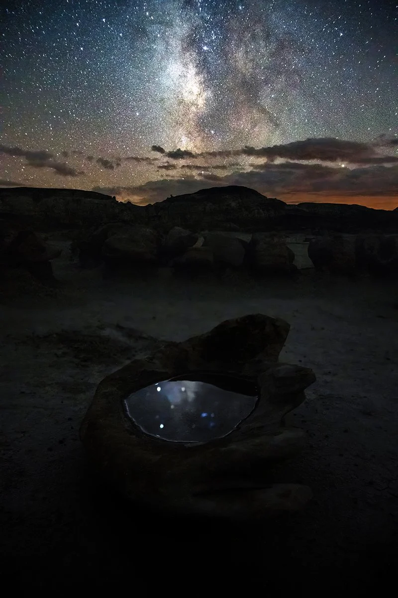 A starry night sky with the Milky Way visible, over a rocky landscape with a large rock containing a puddle of water reflecting the stars.