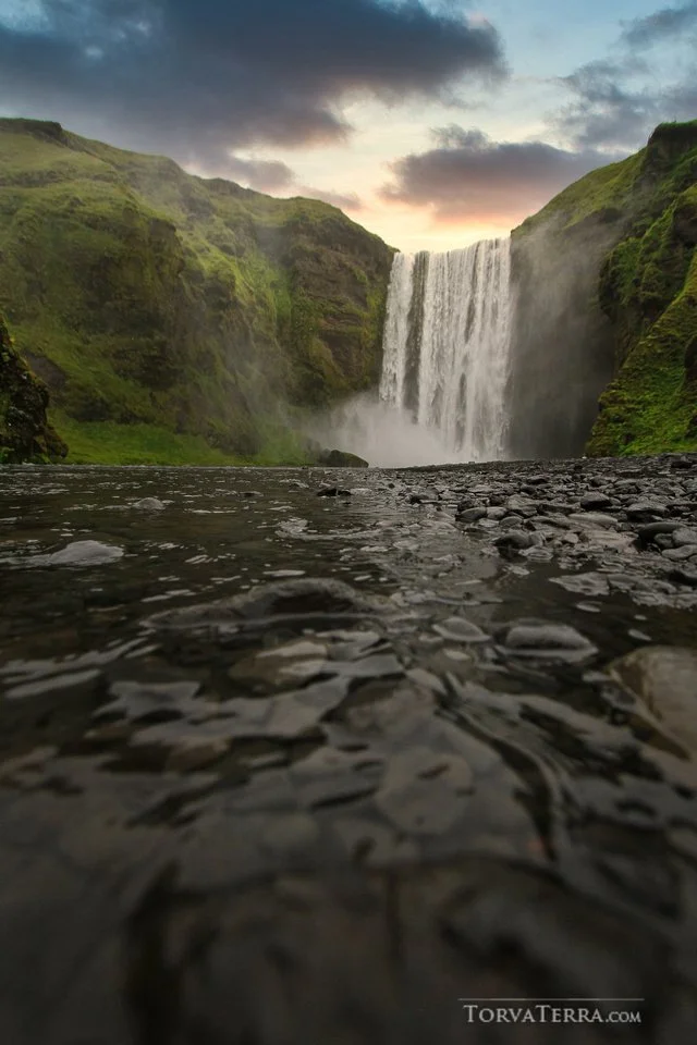 A waterfall cascading down from a cliff into a river with dark rocks, lush green hills on either side, and a cloudy sky at sunset or sunrise.