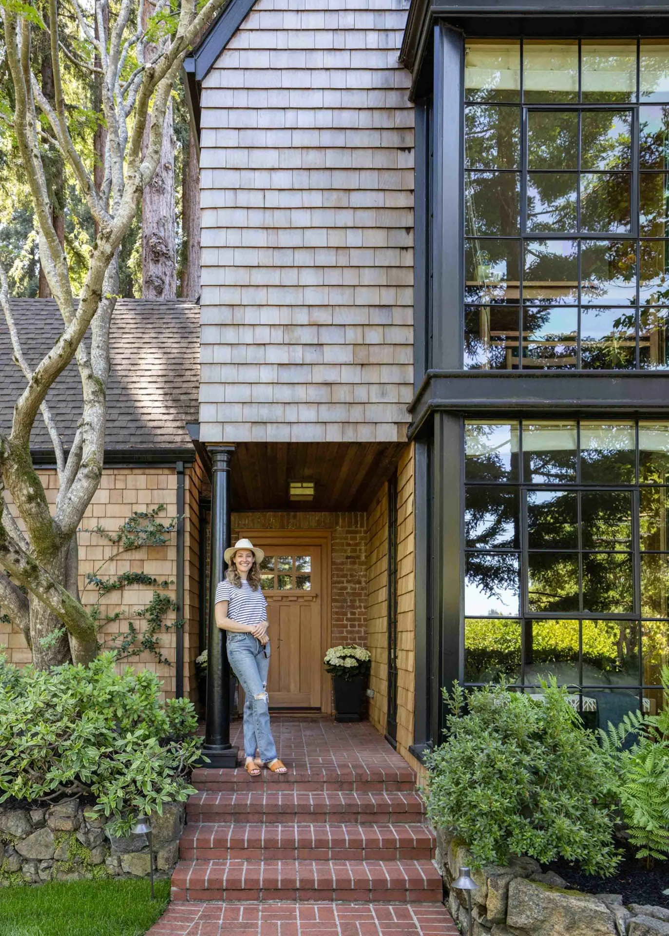 A woman standing on red brick steps outside a house with wood siding, a large window, and lush green plants nearby.