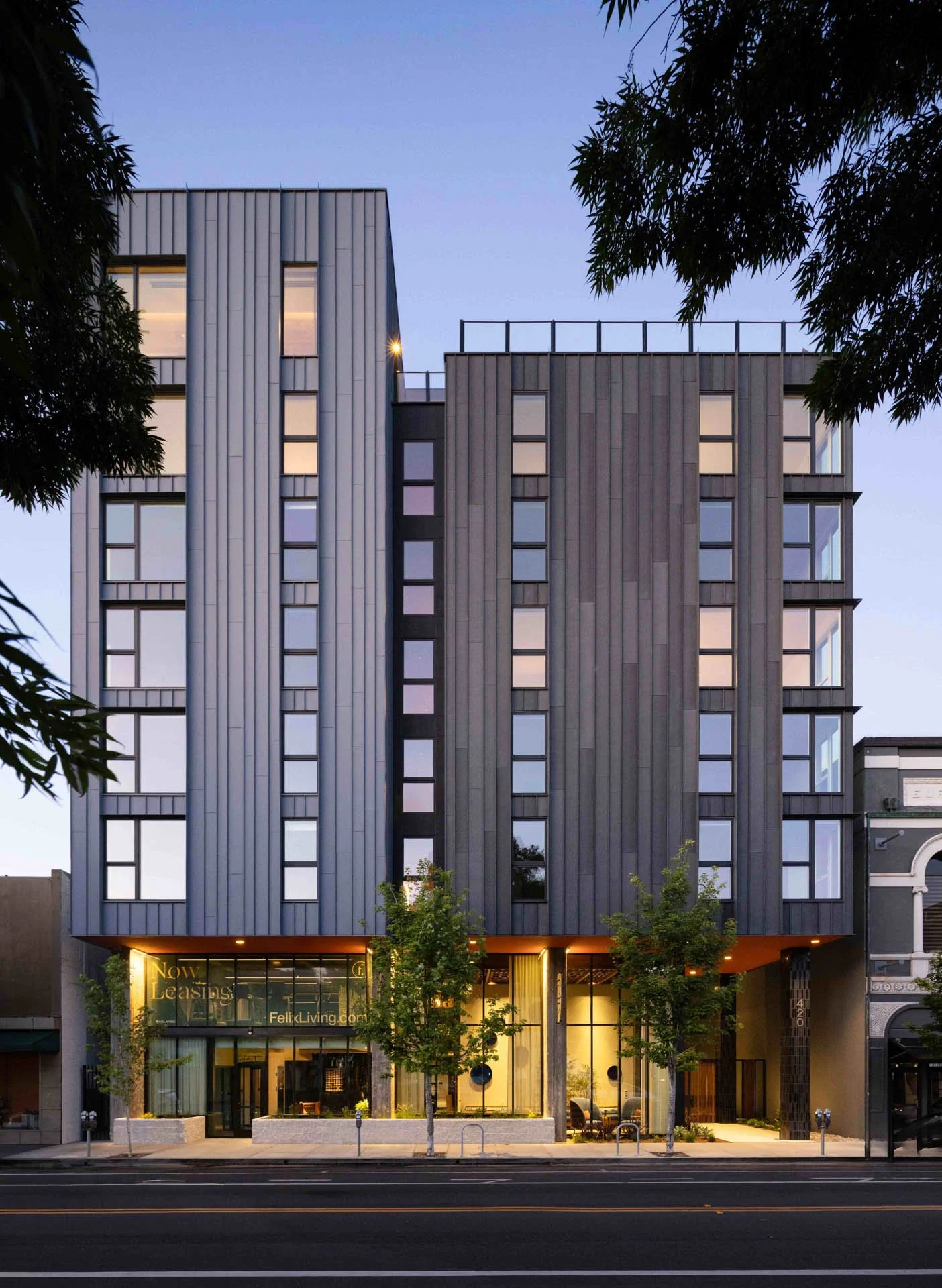 Modern multi-story building with large glass windows and trees at street level, illuminated by exterior lighting at dusk.