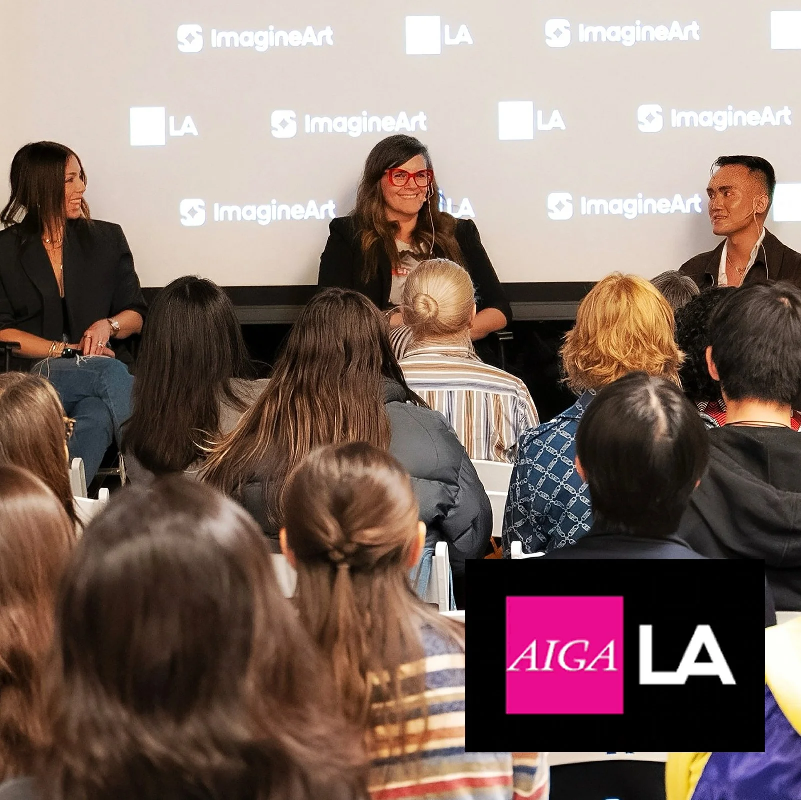 Panel discussion with three women on stage at an event, the woman in the center wearing red glasses and speaking to an audience, with a large screen behind them displaying the 'ImagineArt' logo.