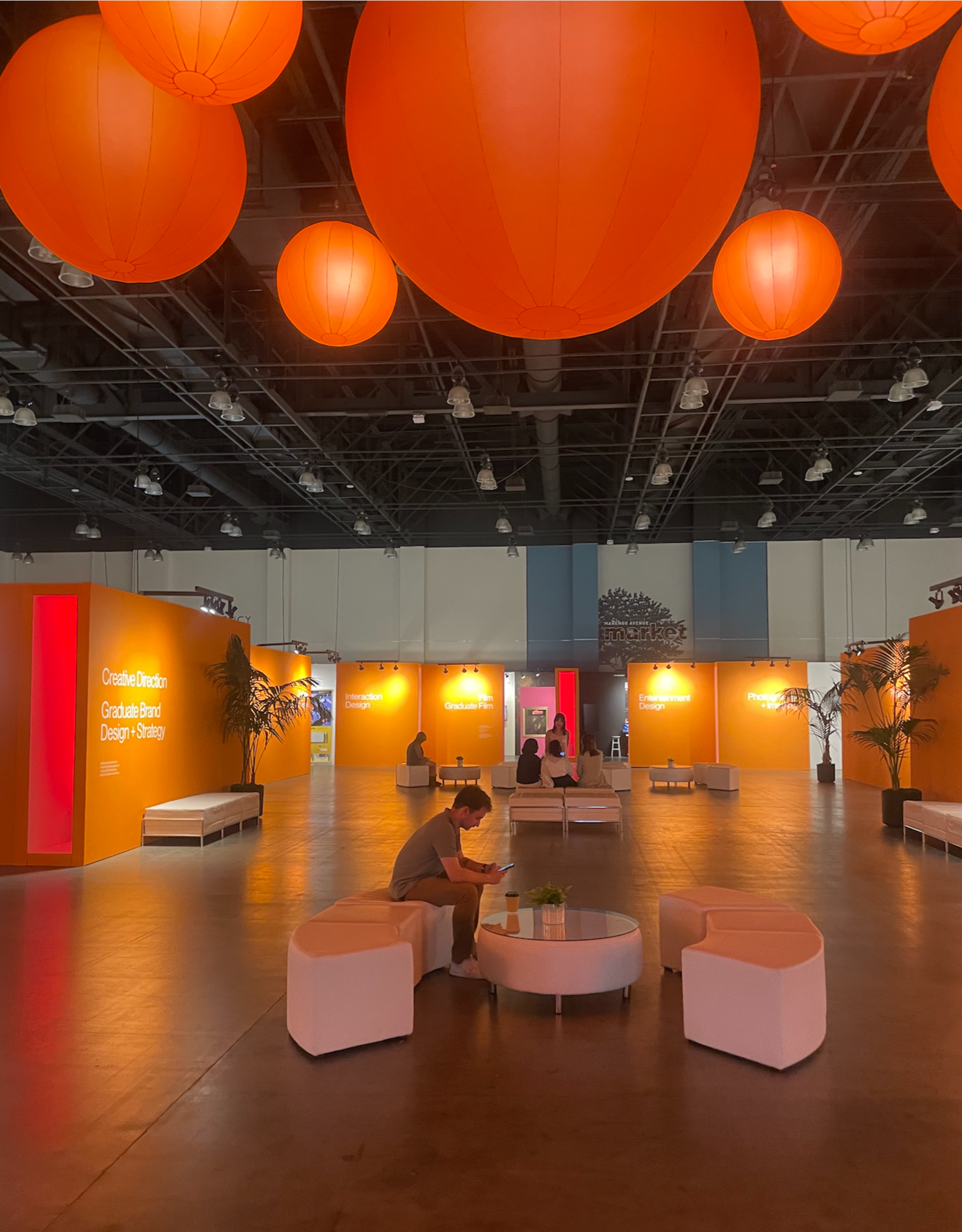 Indoor event space with orange paper lanterns on the ceiling, orange display walls with text, white seating areas, and a few people sitting and using their phones.