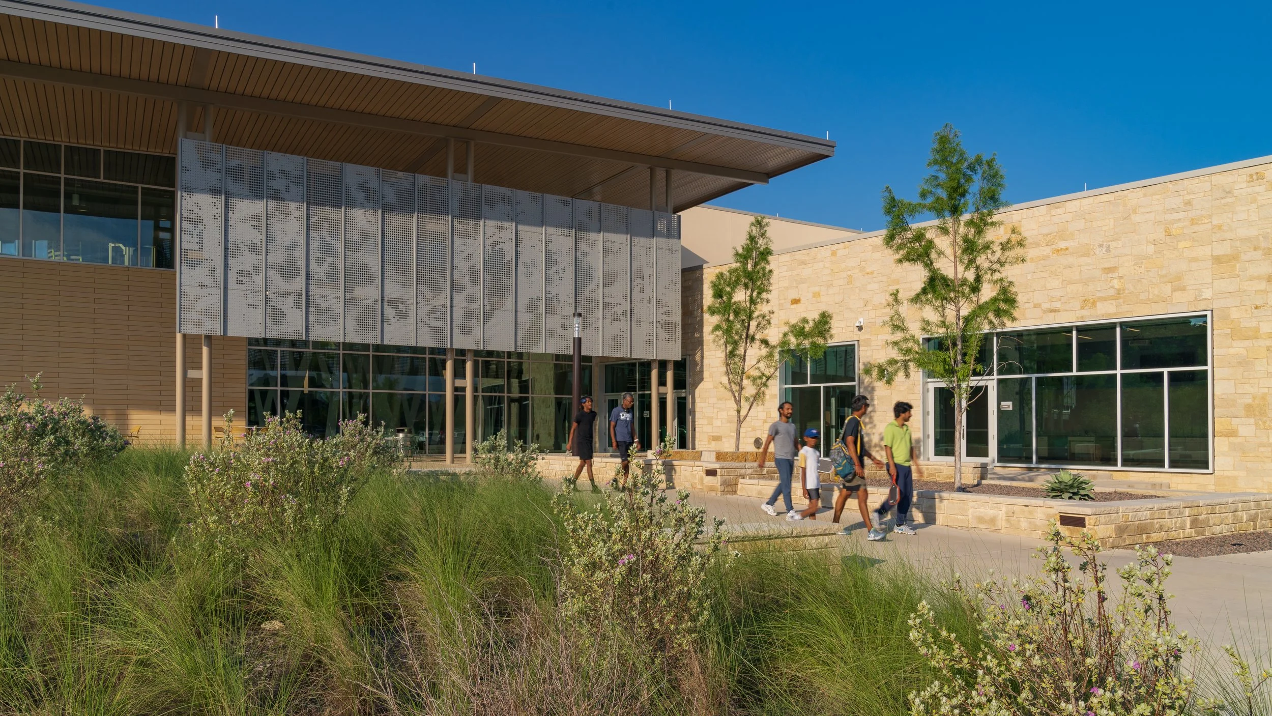 A modern building with large windows, a stone and wood exterior, and a metal decorative panel on the front. Several people are walking on the sidewalk in front of the building, with trees and bushes nearby under a clear blue sky.