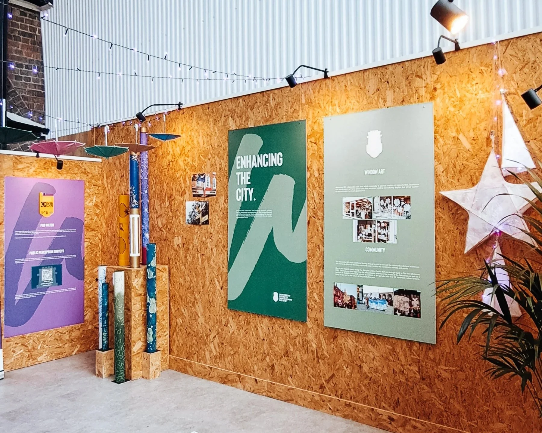 An art exhibit with informational posters mounted on a wooden wall, including one large green poster titled "Enhancing the City", smaller photos and text, decorative star and paper lanterns, and assorted cylindrical objects stacked on wooden blocks.