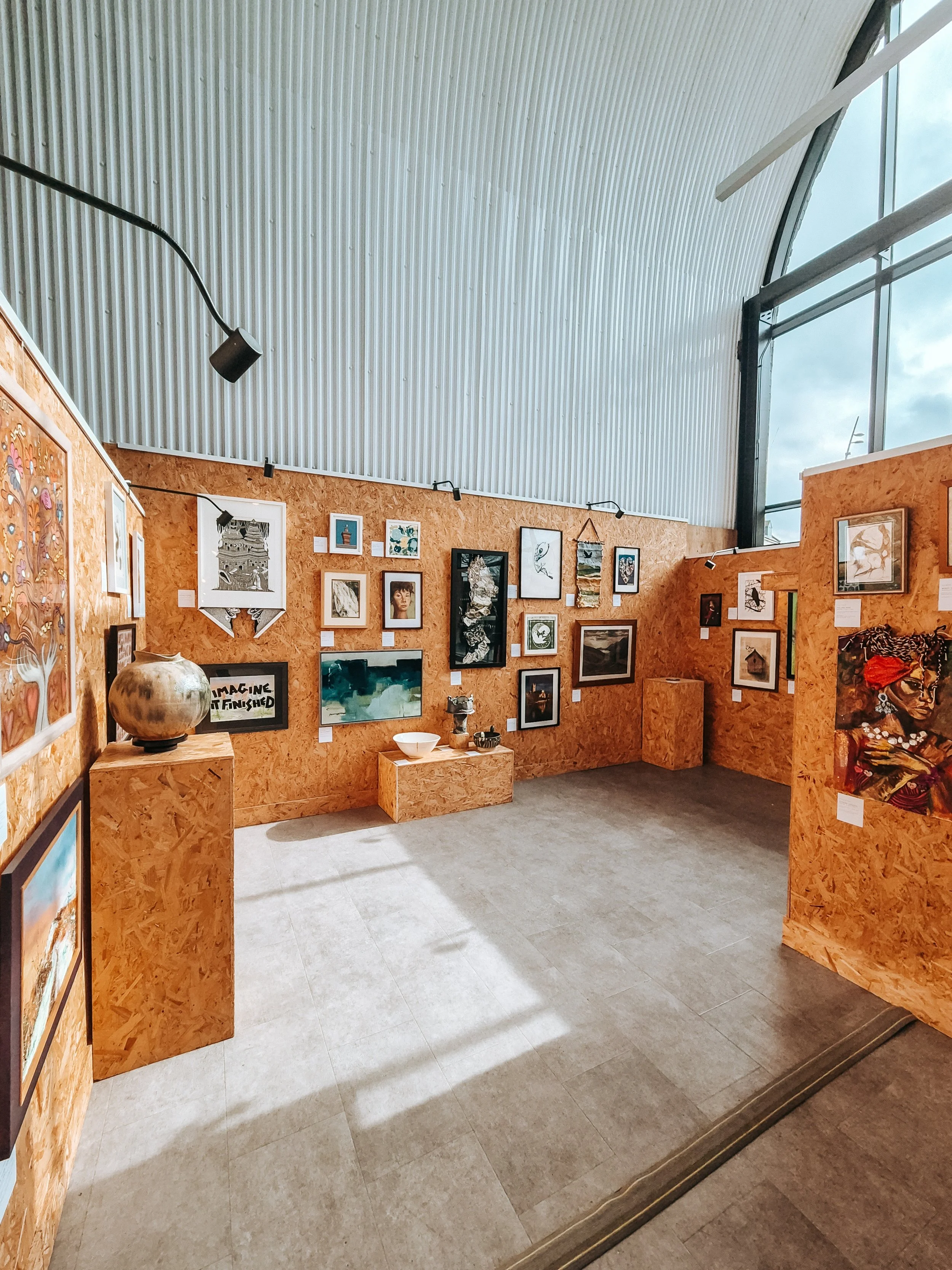 An art gallery with framed paintings and photographs on orange-colored wooden walls, featuring sculptures and artifacts displayed on pedestals, with natural light coming through large windows and track lighting overhead.