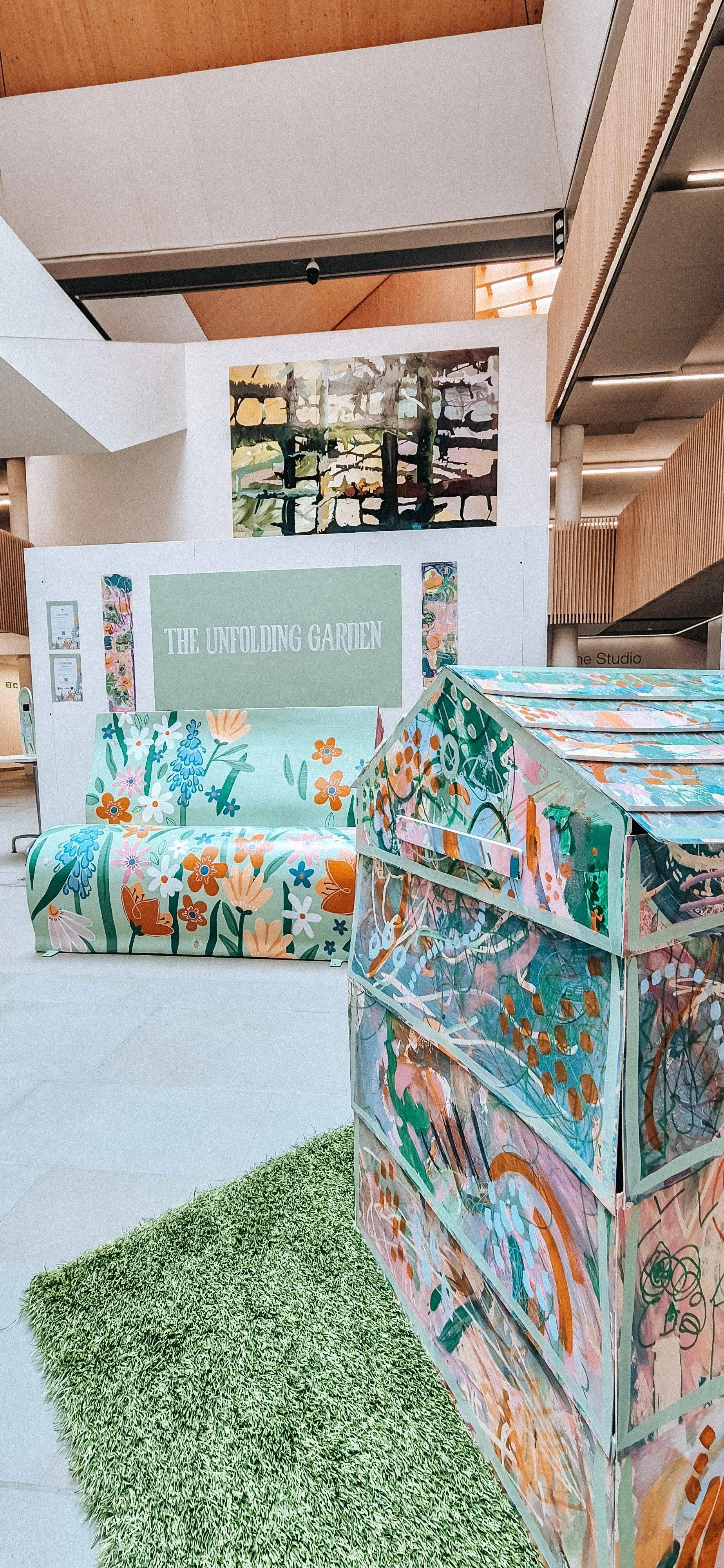Colorful art installation titled 'The Unfolding Garden' featuring a floral patterned sofa, an abstract painting on the wall, a multicolored art display in the foreground, a green shaggy rug, and wooden panels in a modern interior setting.