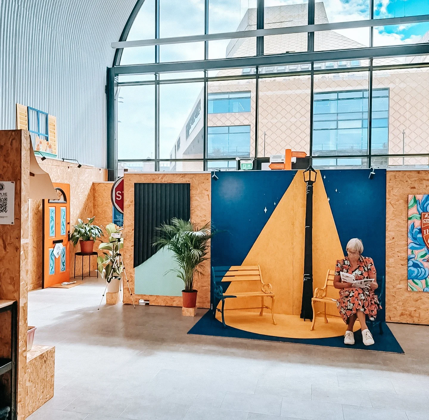 A woman with short grey hair, wearing glasses and a patterned dress, sits on a yellow bench, reading a book, in a modern indoor space with large windows, plants, and colorful wall art.