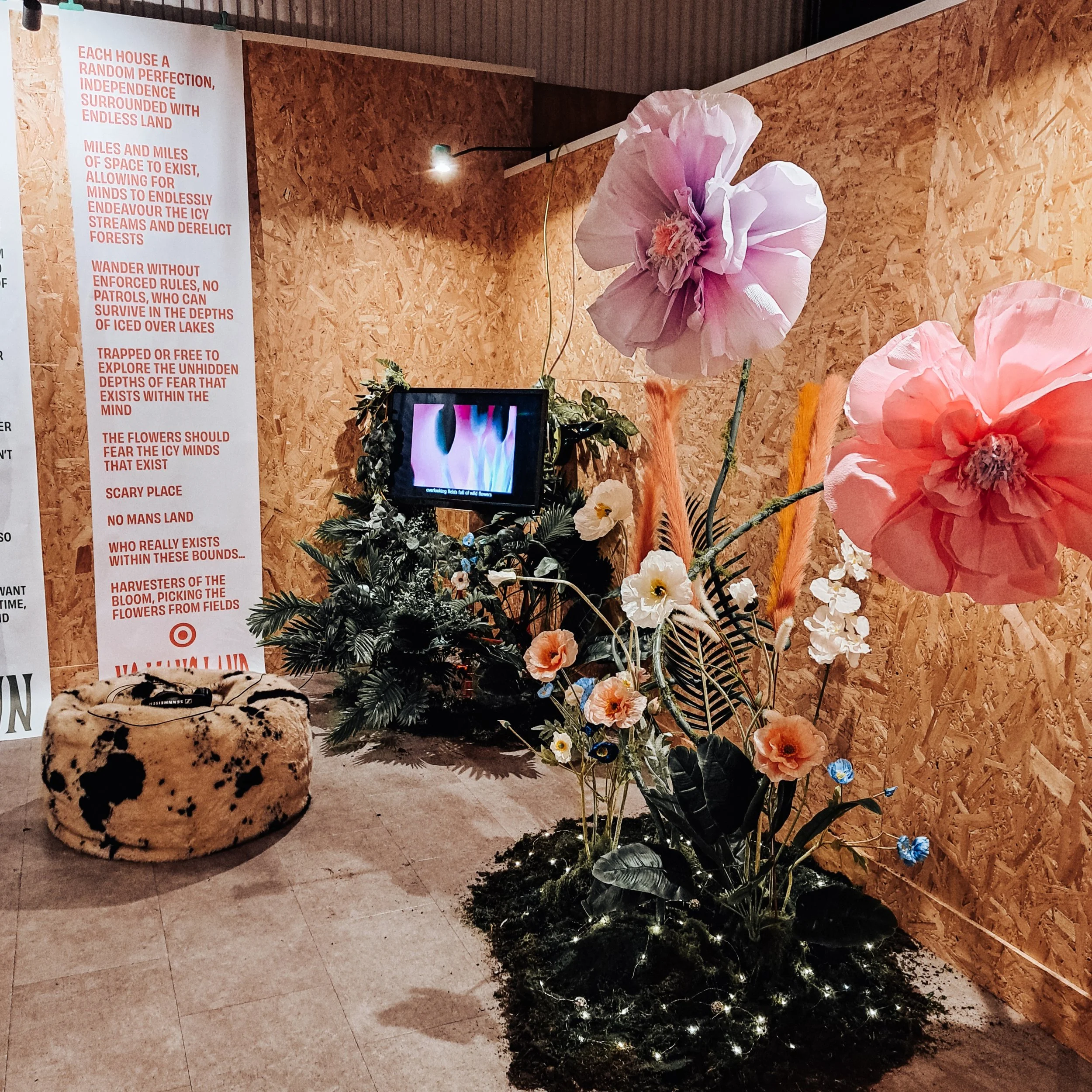 An art installation featuring large, colorful paper flowers, including pink and orange poppies, with a small TV displaying animated flames. There is a cow-patterned round stool and a wall with a red and white motivational poster text on the left. The background is a textured wood wall.